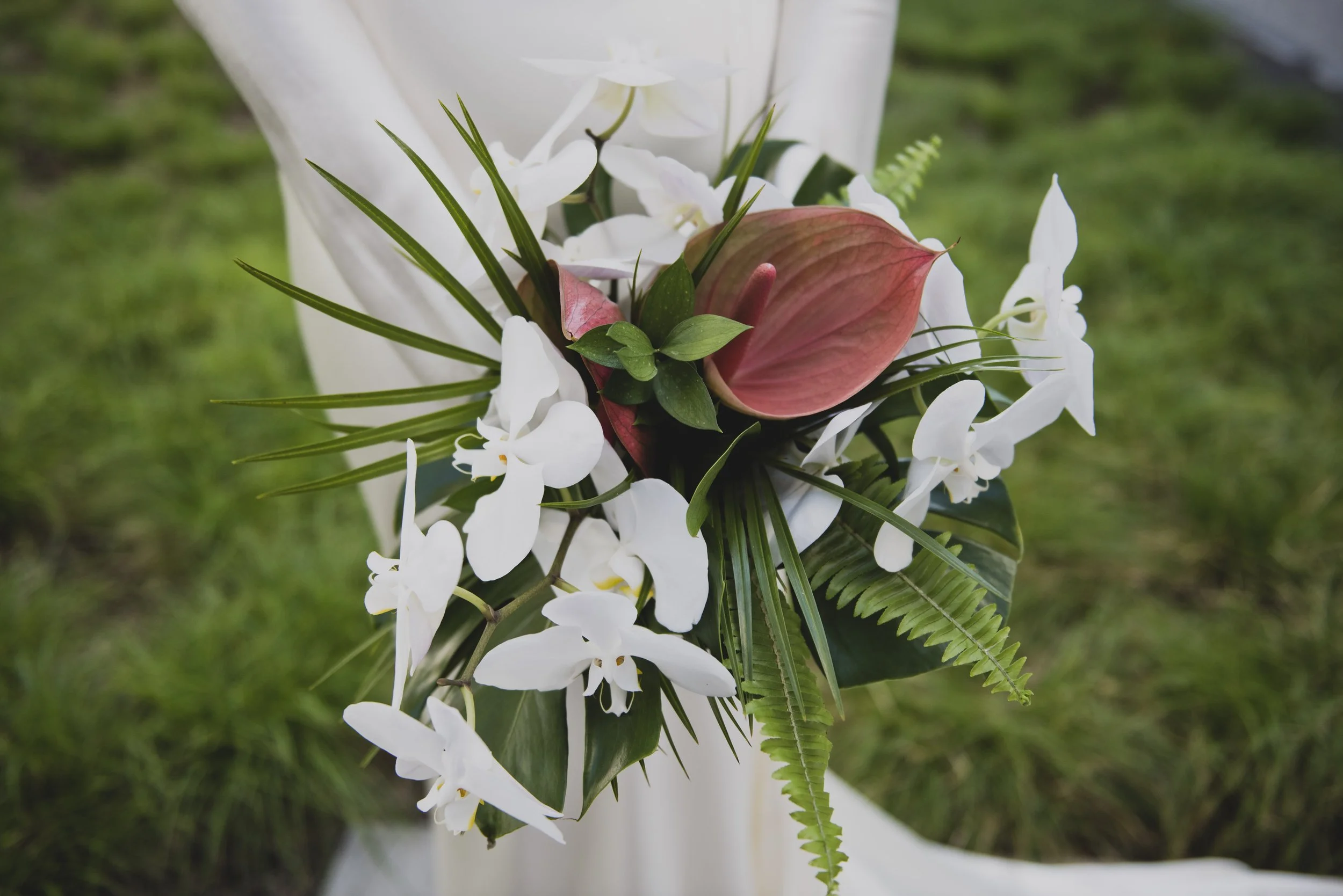 Close-up of a contemporary tropical bridal bouquet with white orchids, blush anthuriums, monstera leaves, palms, and ferns for a modern wedding celebration.