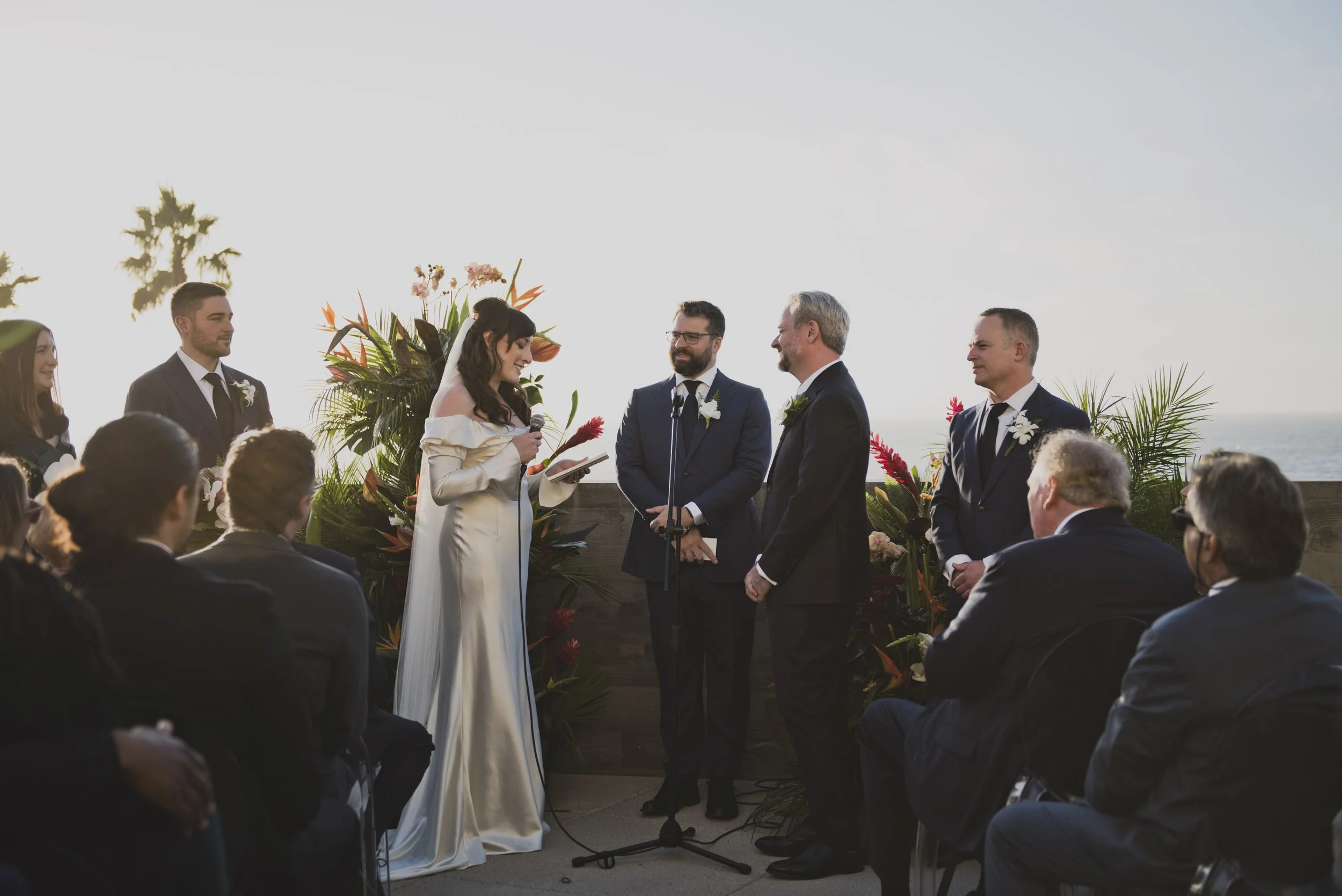 Bride reading her handwritten wedding vows to the groom during an outdoor oceanfront wedding ceremony with family and friends in attendance.
