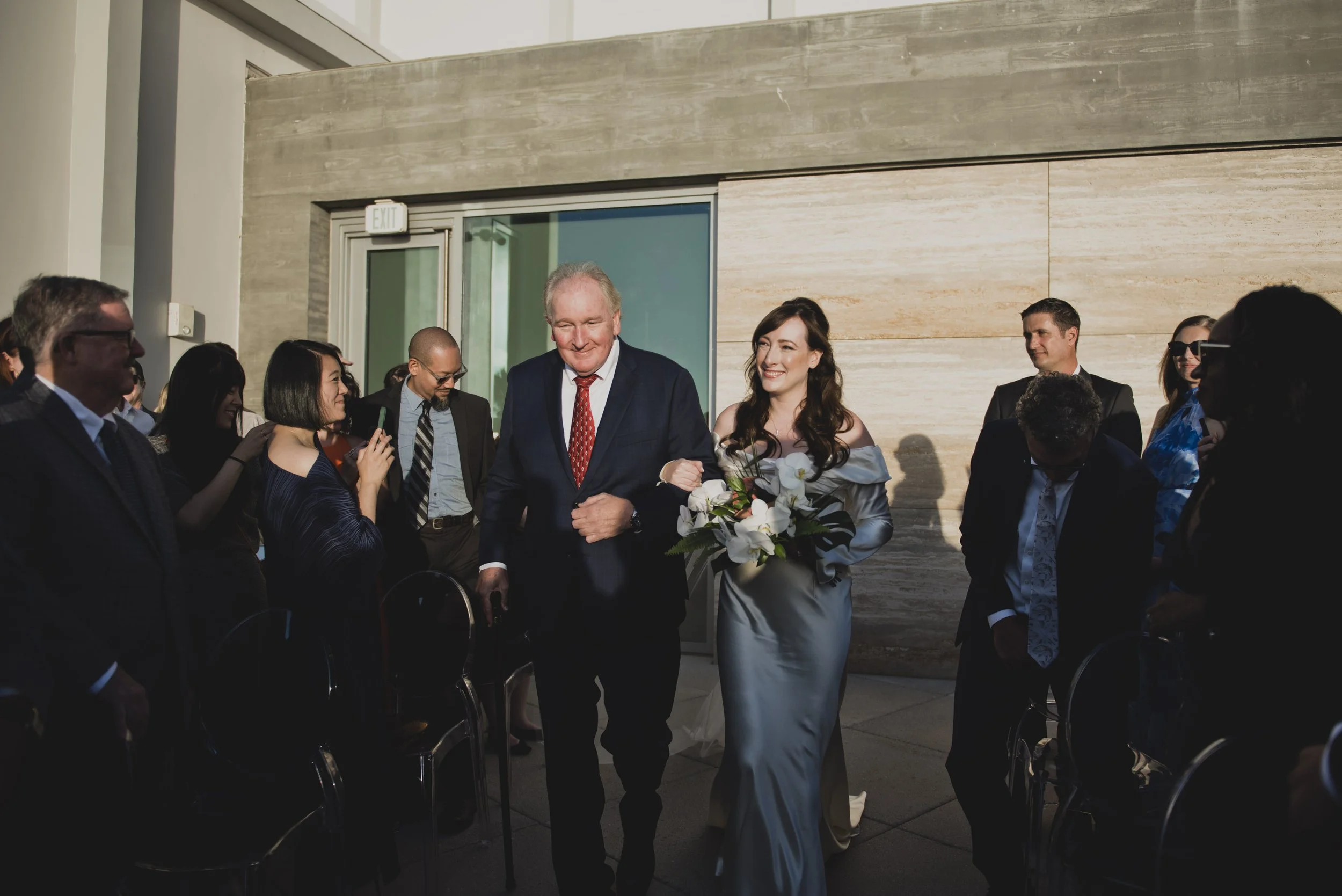 Bride walking down the aisle with her father while carrying a tropical orchid bouquet at an oceanfront wedding ceremony.