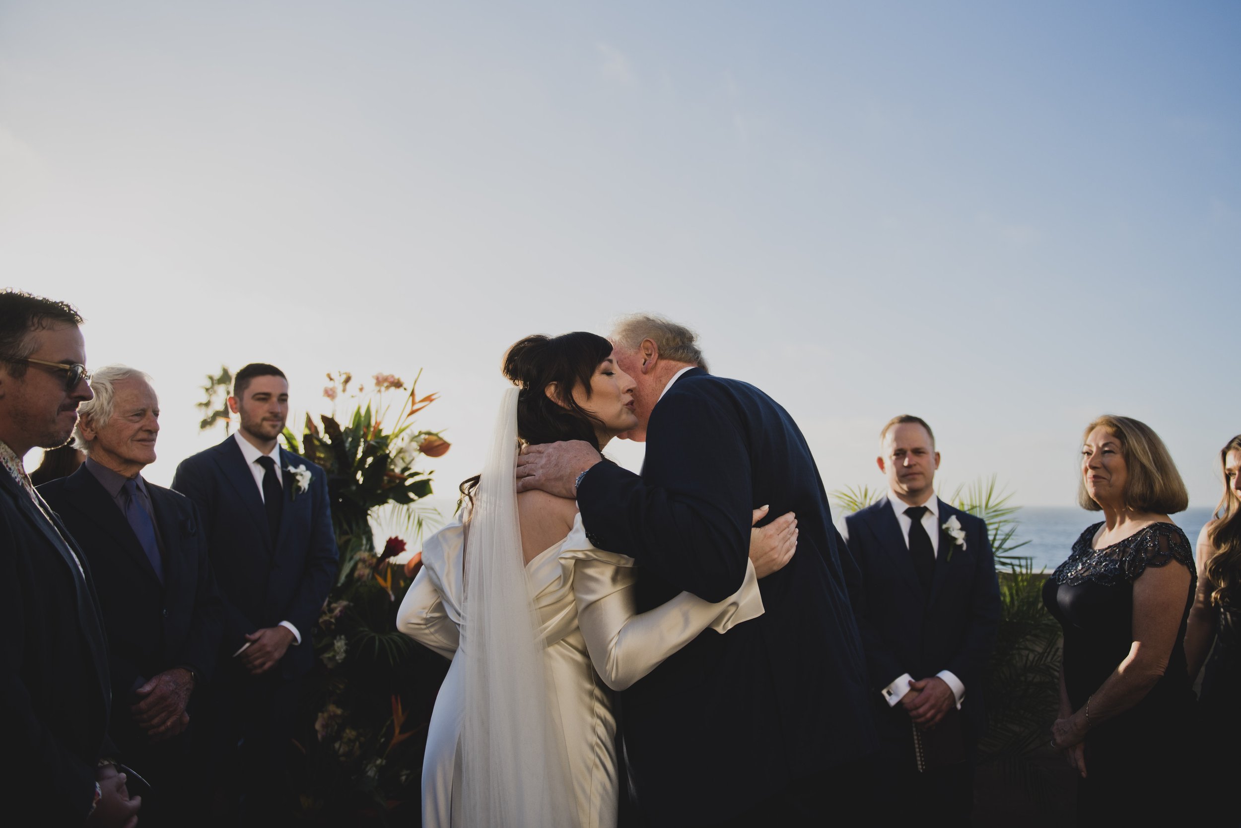 Father embracing the bride during an emotional moment at an outdoor oceanfront wedding ceremony before giving her away.