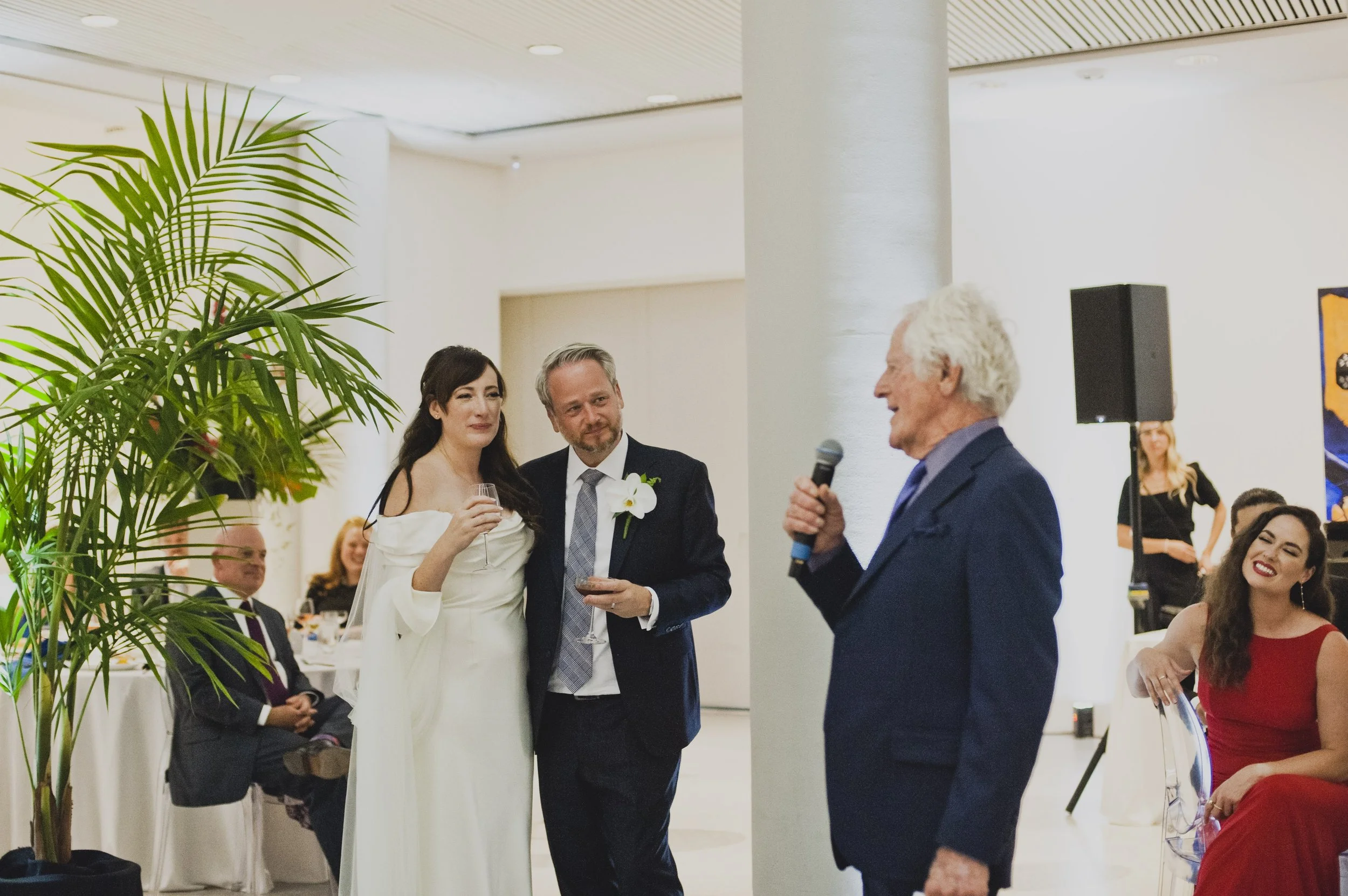 Bride and groom listening to a heartfelt wedding reception toast while holding wine glasses as a family member gives a speech.