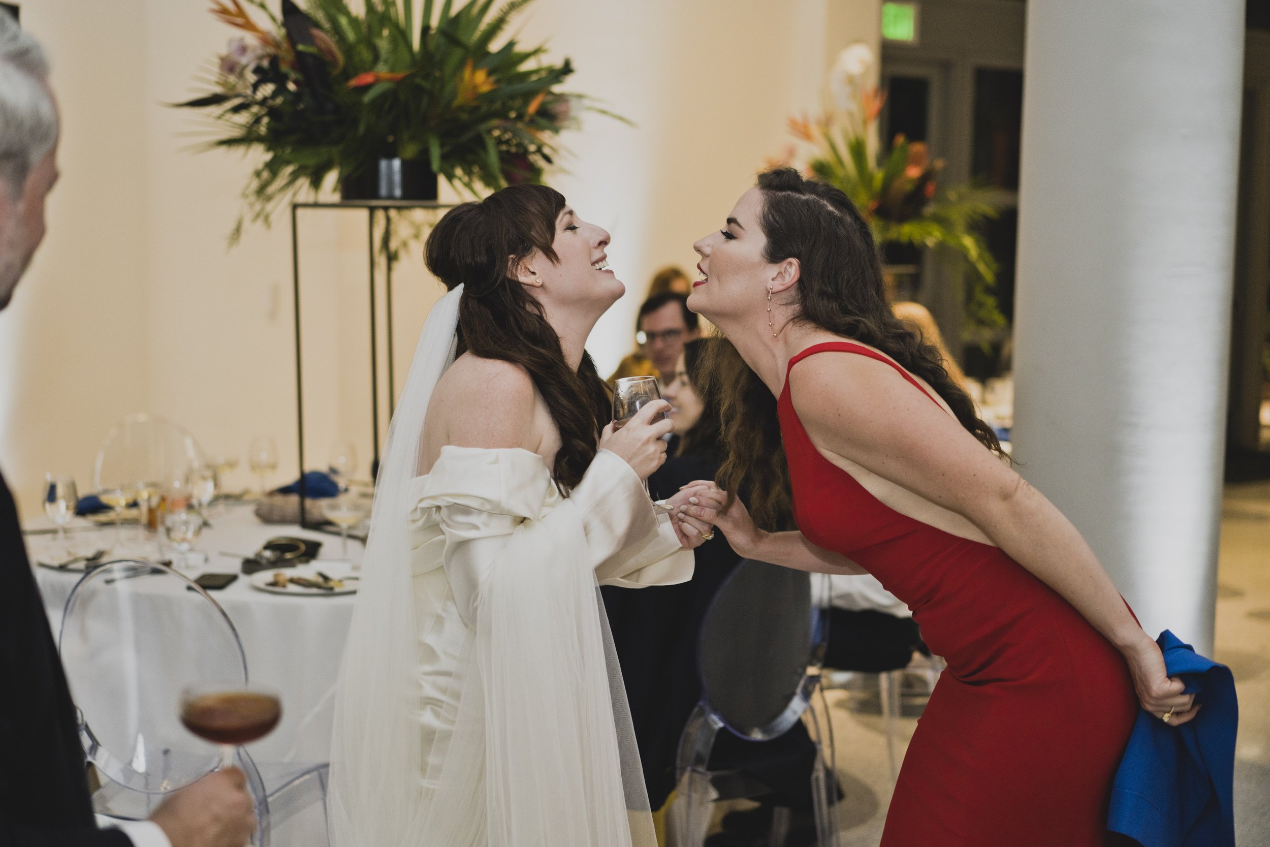 Bride warmly greeting a wedding guest during the reception inside a contemporary museum venue surrounded by elegant tropical floral décor.