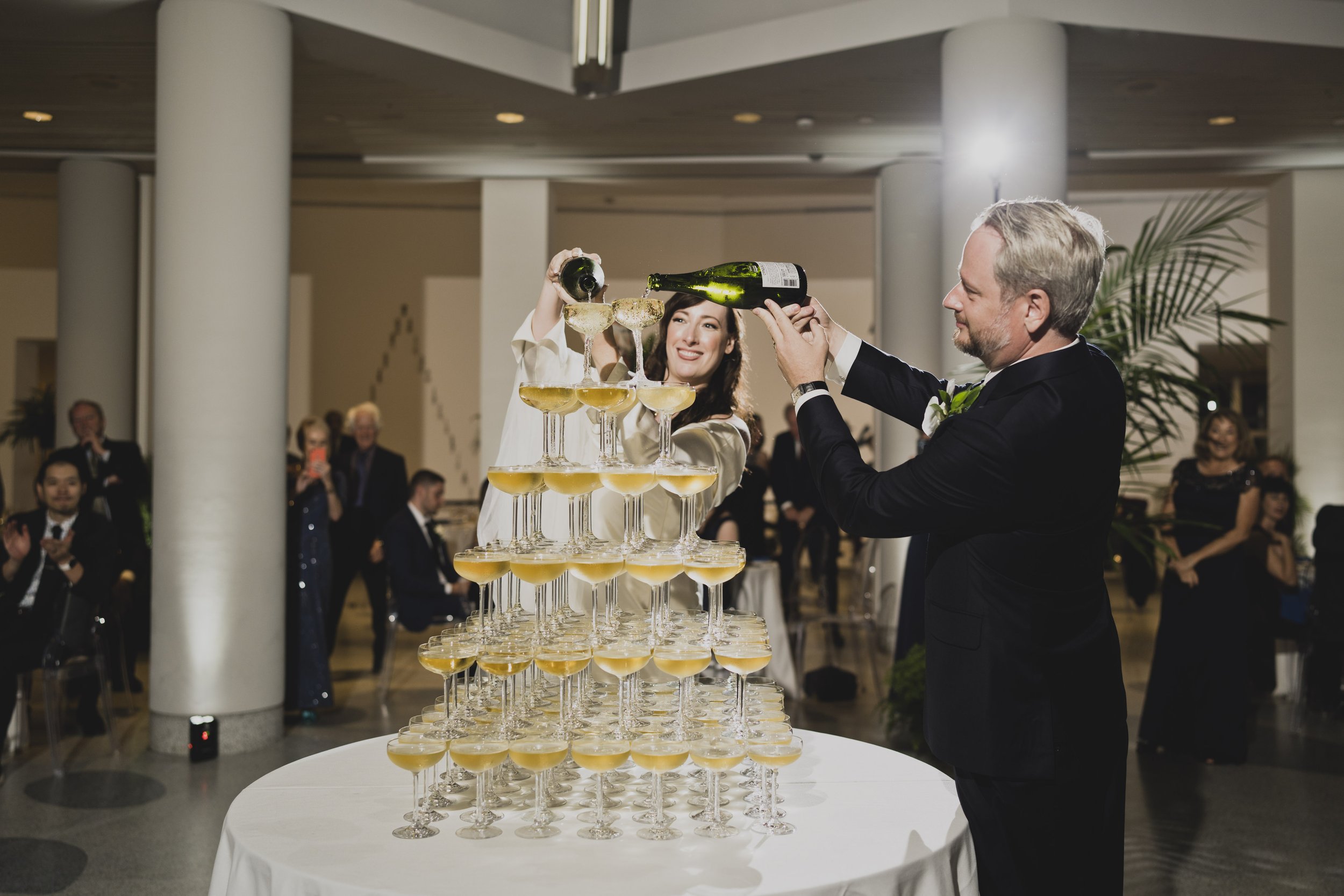 Bride and groom pouring champagne together into a champagne tower while guests gather to celebrate during their modern museum wedding reception.