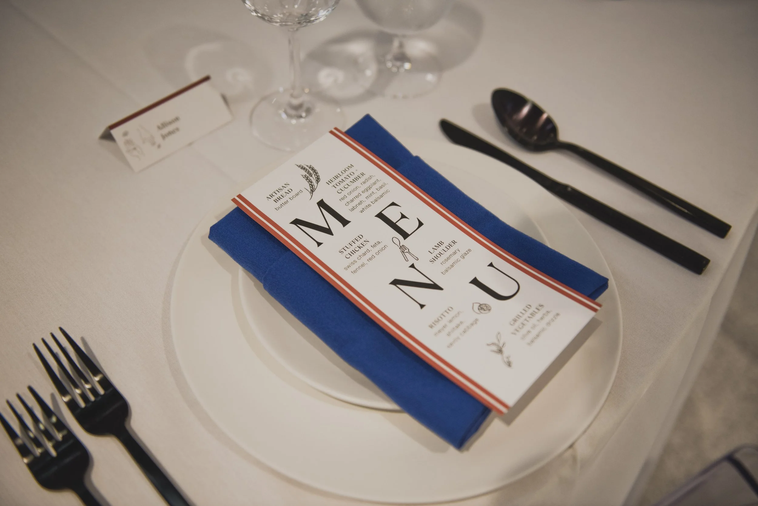 Wedding reception place setting featuring a custom printed menu, royal blue linen napkin, black flatware, and white dinnerware on a modern reception table.