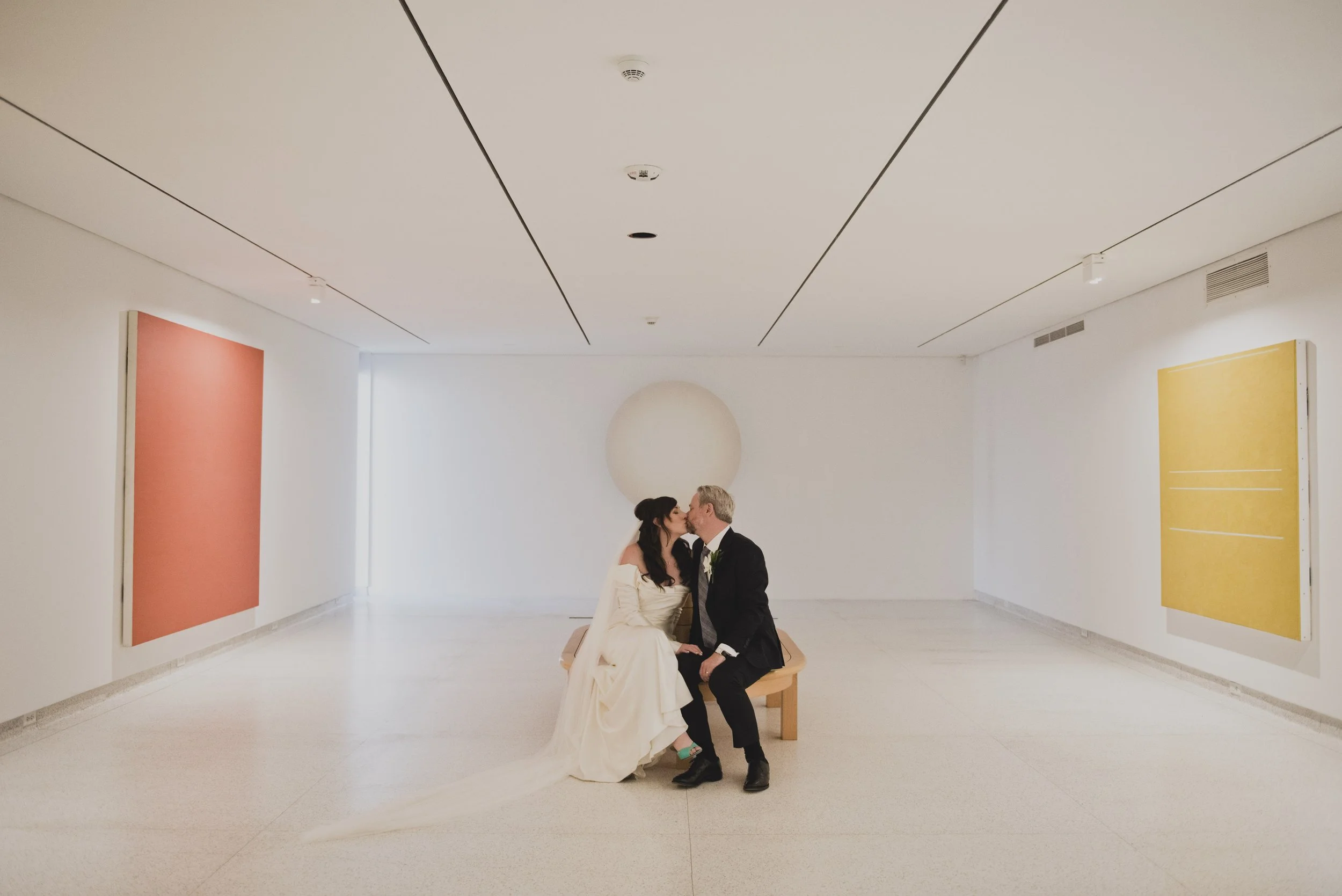 Bride and groom sharing a kiss while seated on a gallery bench between colorful modern art installations inside a contemporary art museum.