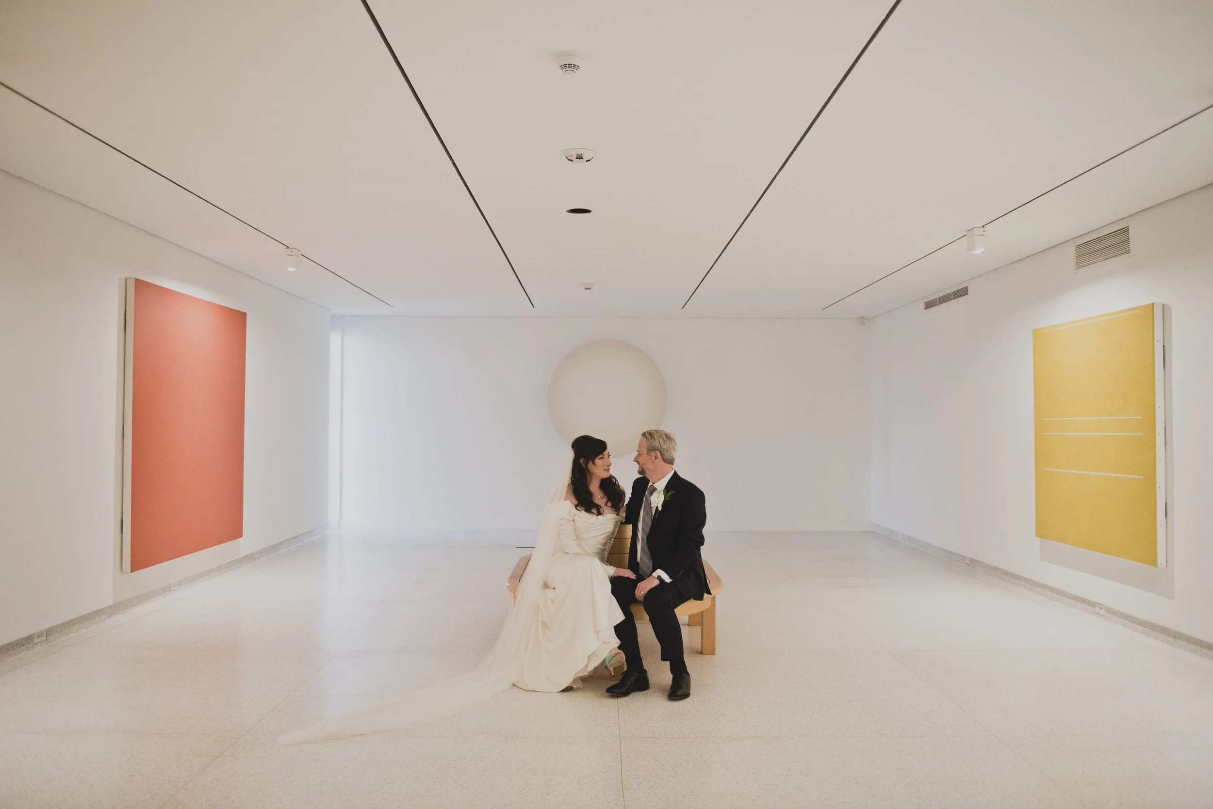 Bride and groom seated together on a gallery bench beneath minimalist artwork, sharing a quiet moment inside a contemporary art museum after their wedding ceremony.