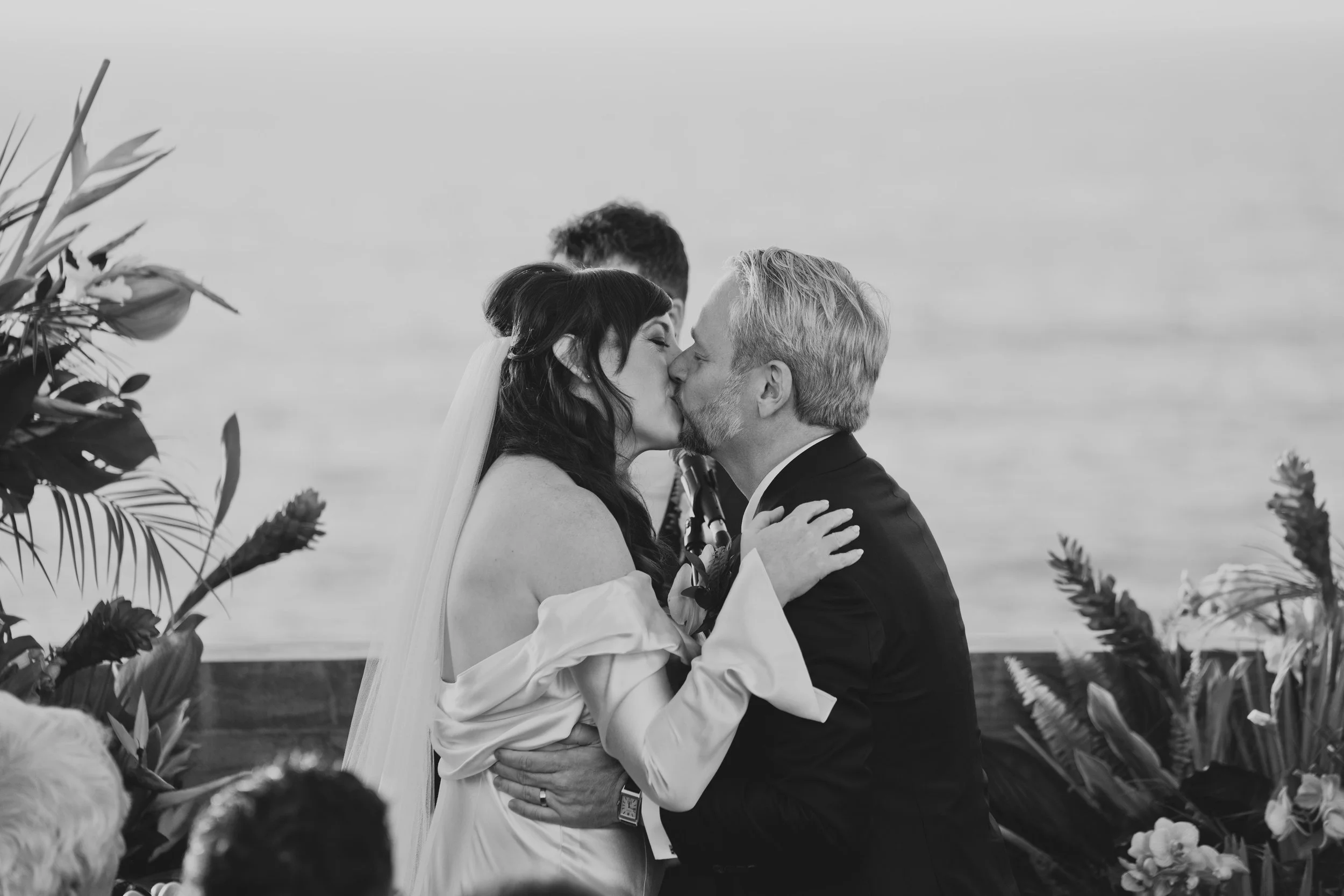 Bride and groom sharing their first kiss as newlyweds during a modern oceanfront wedding ceremony with tropical floral arrangements in the background.