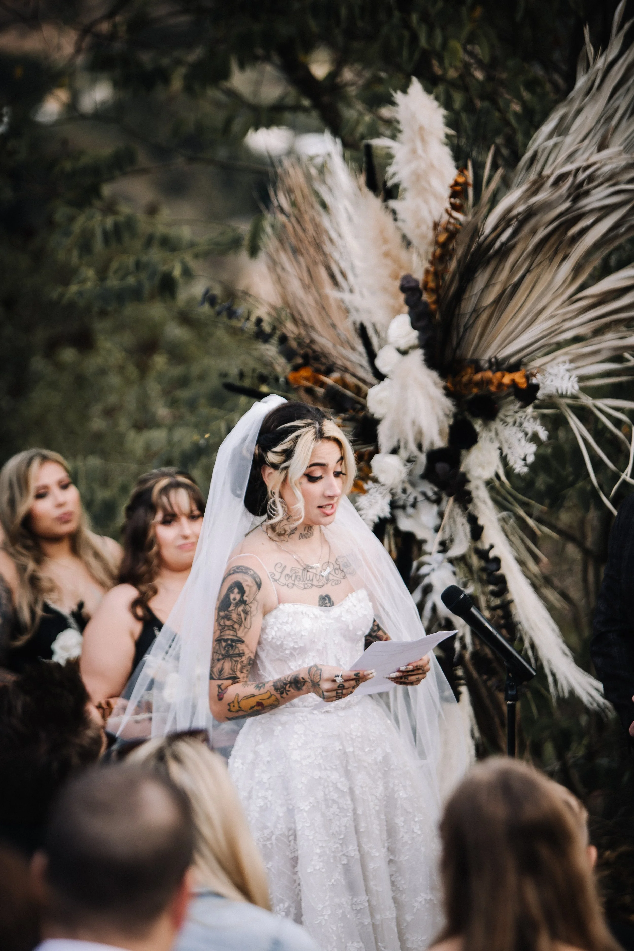Bride reads her handwritten vows during an outdoor Halloween wedding ceremony beneath a dramatic dried floral installation with black and white accents.