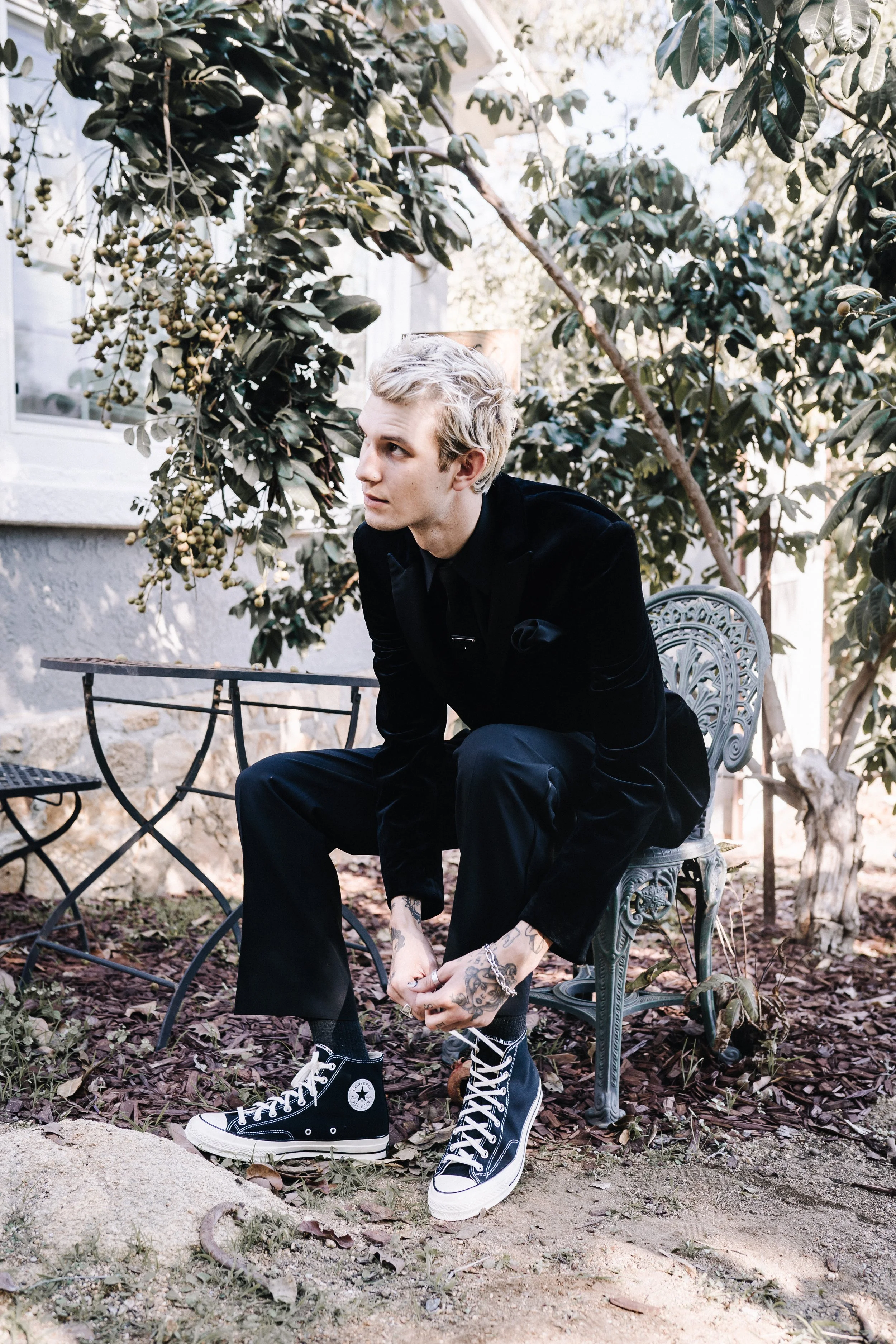 Groom in a black velvet suit and Converse sneakers seated outdoors before an alternative Halloween wedding ceremony.
