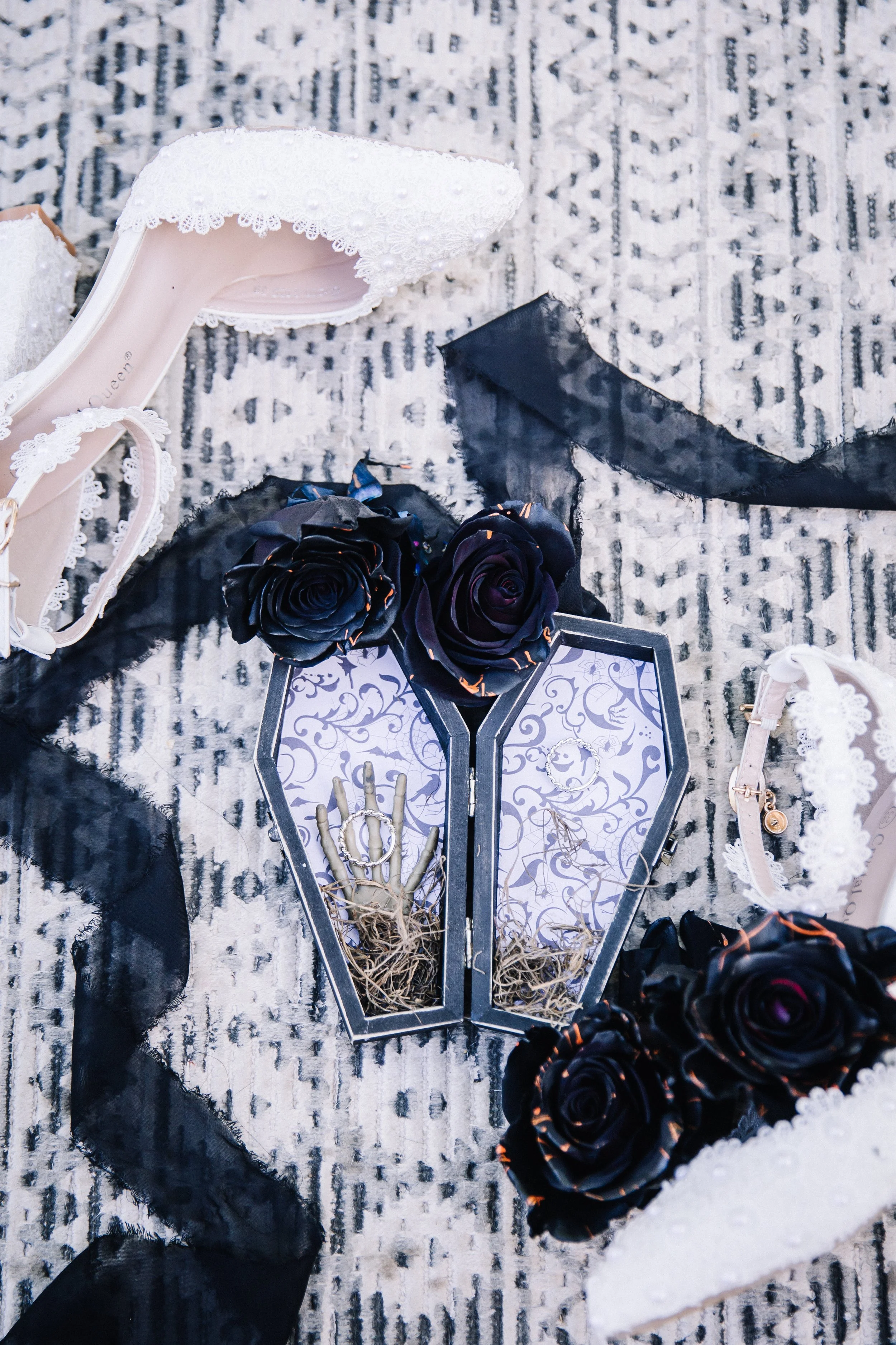 Halloween wedding detail flat lay featuring lace bridal heels, black roses, a coffin ring box, skeleton ring holder, black ribbon, and wedding accessories.