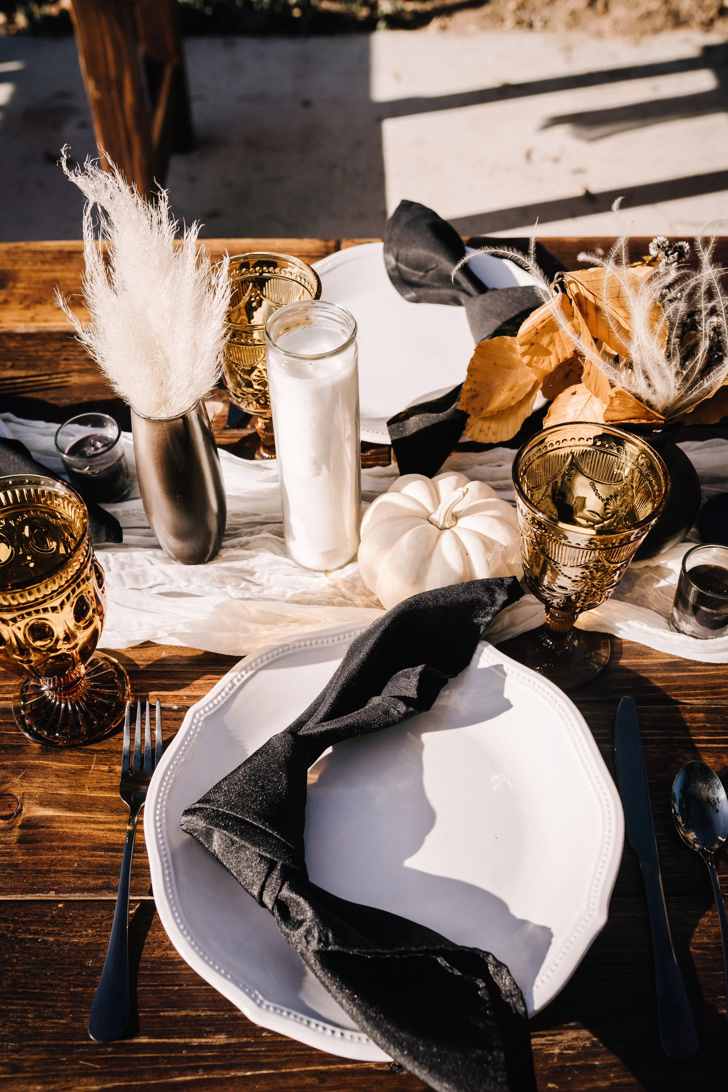 Elegant Halloween wedding reception table with white dinnerware, black napkins, amber goblets, candles, pampas grass, and white pumpkins on a rustic wood table.