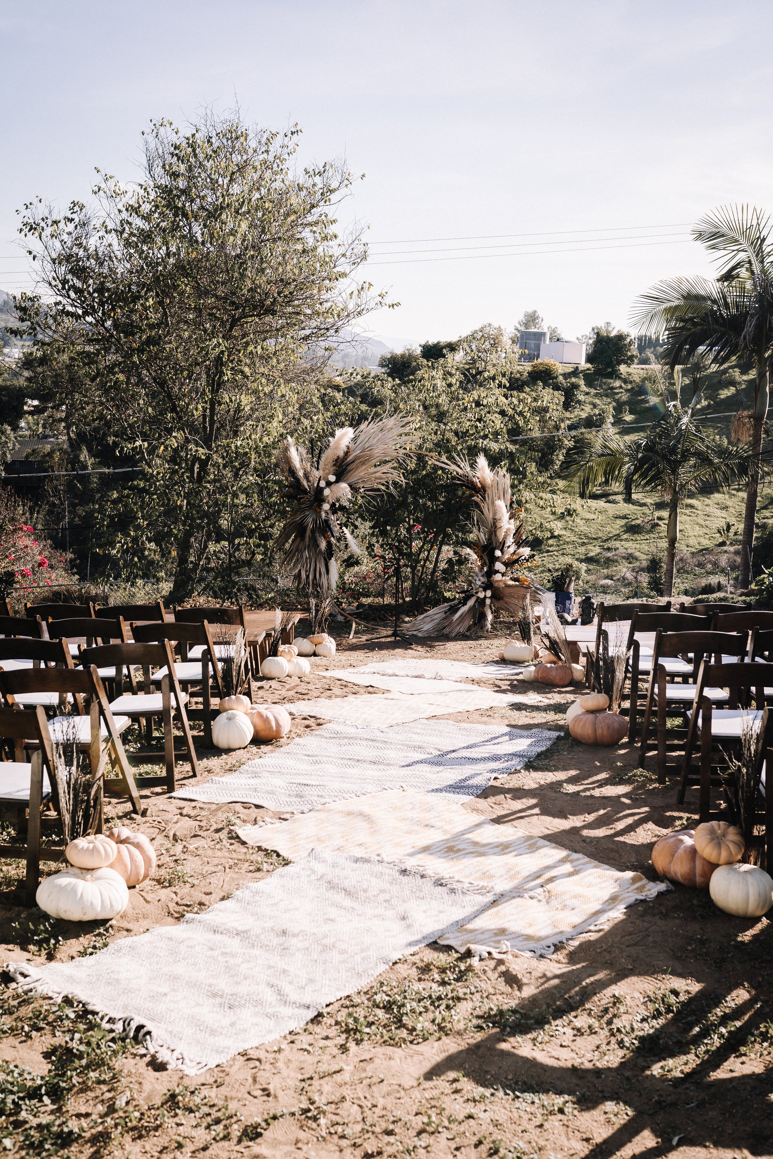 Outdoor Halloween wedding ceremony setup with layered aisle rugs, pampas grass ceremony arches, white pumpkins, and wooden guest seating.