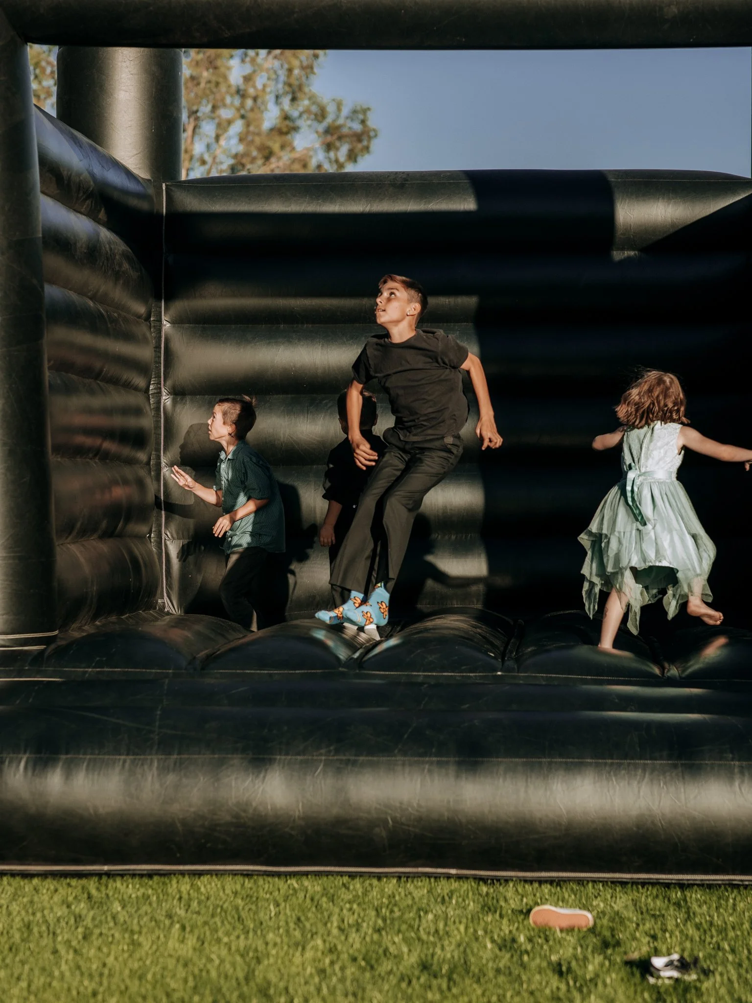Children laughing and jumping together inside a black wedding bounce house during an outdoor wedding reception.