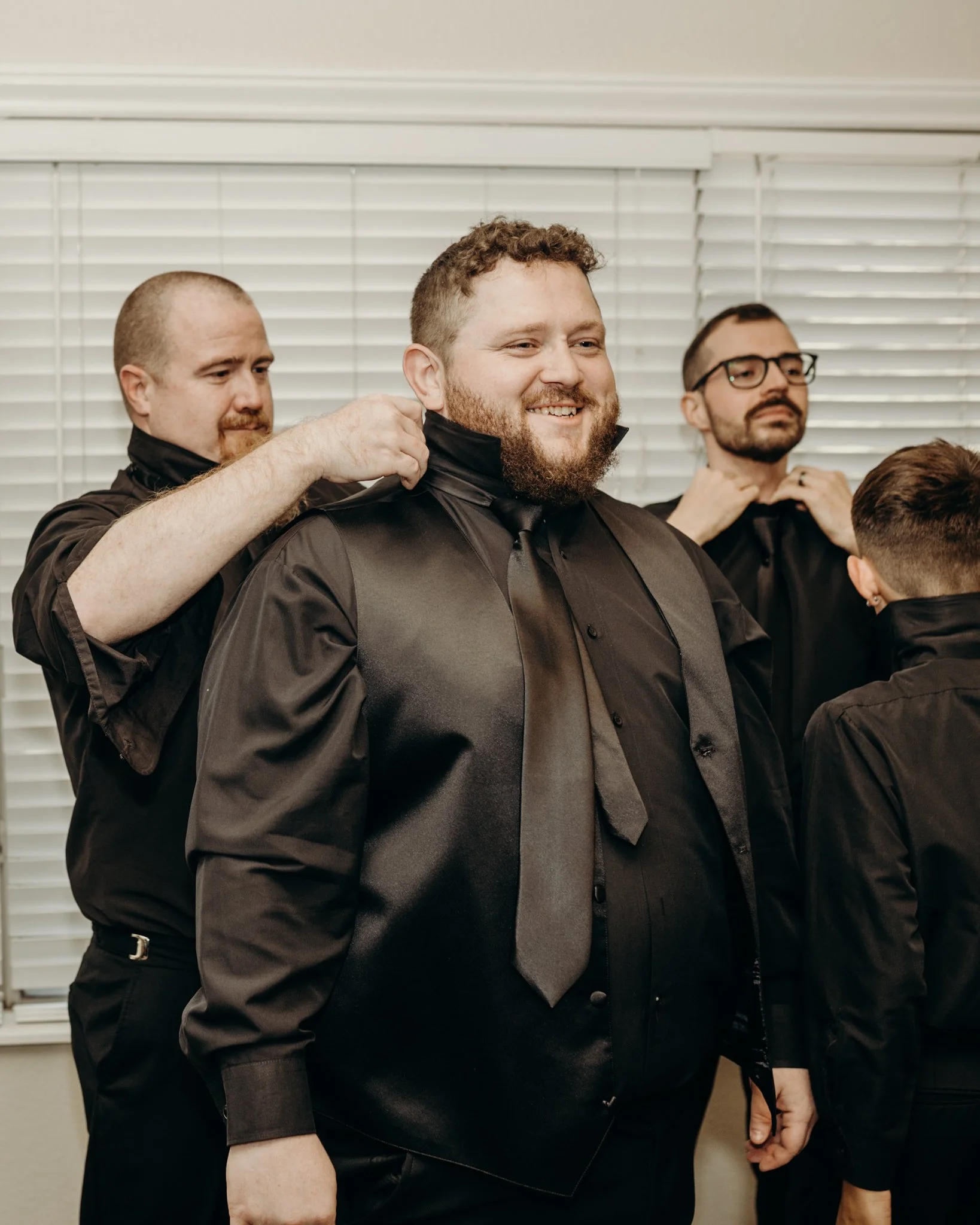 Groom smiling as his groomsmen help straighten his collar and tie while getting ready before the wedding ceremony.