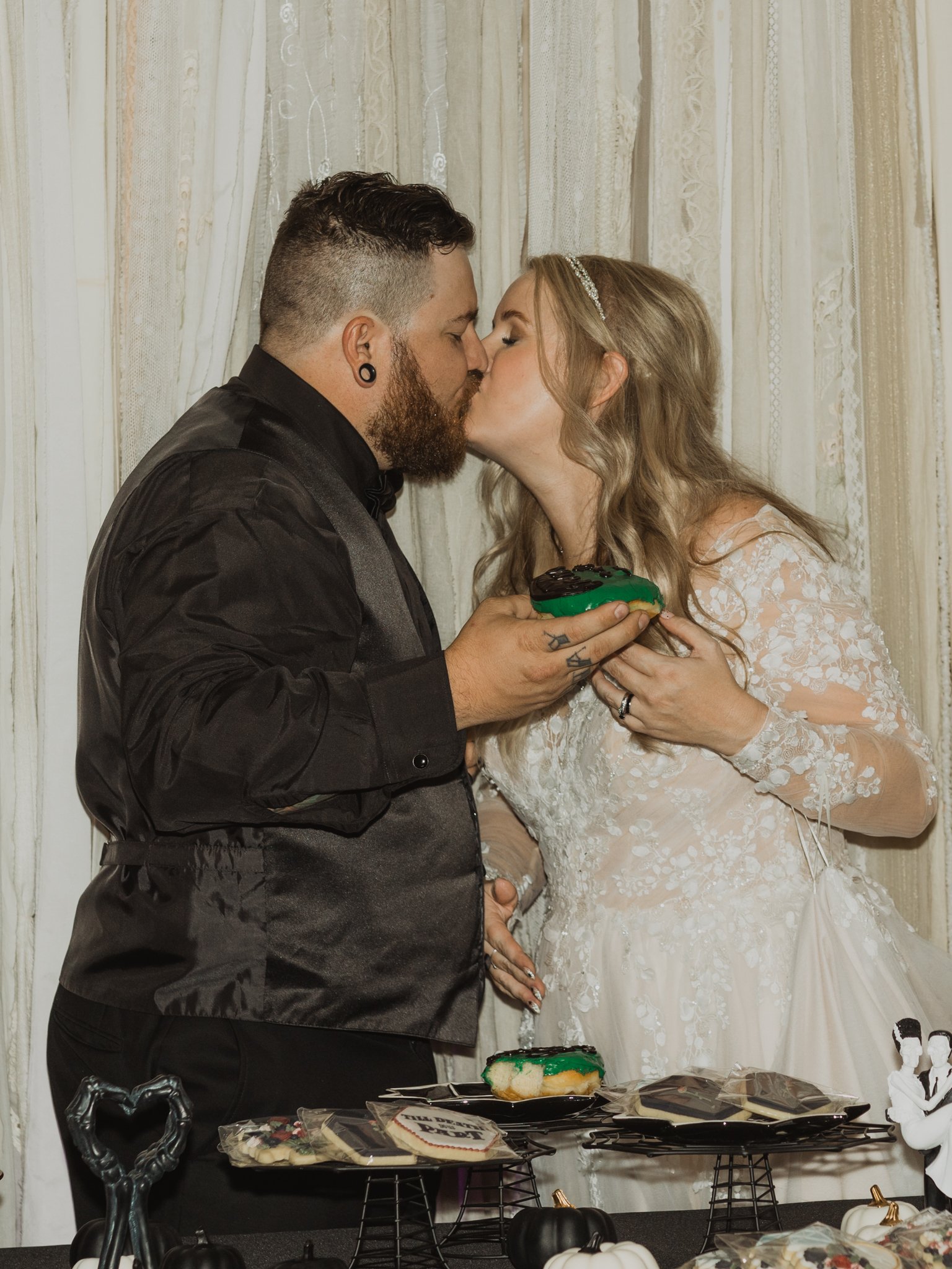 Bride and groom share a kiss while holding a Halloween-themed donut during their unique cake-cutting alternative at the wedding reception.