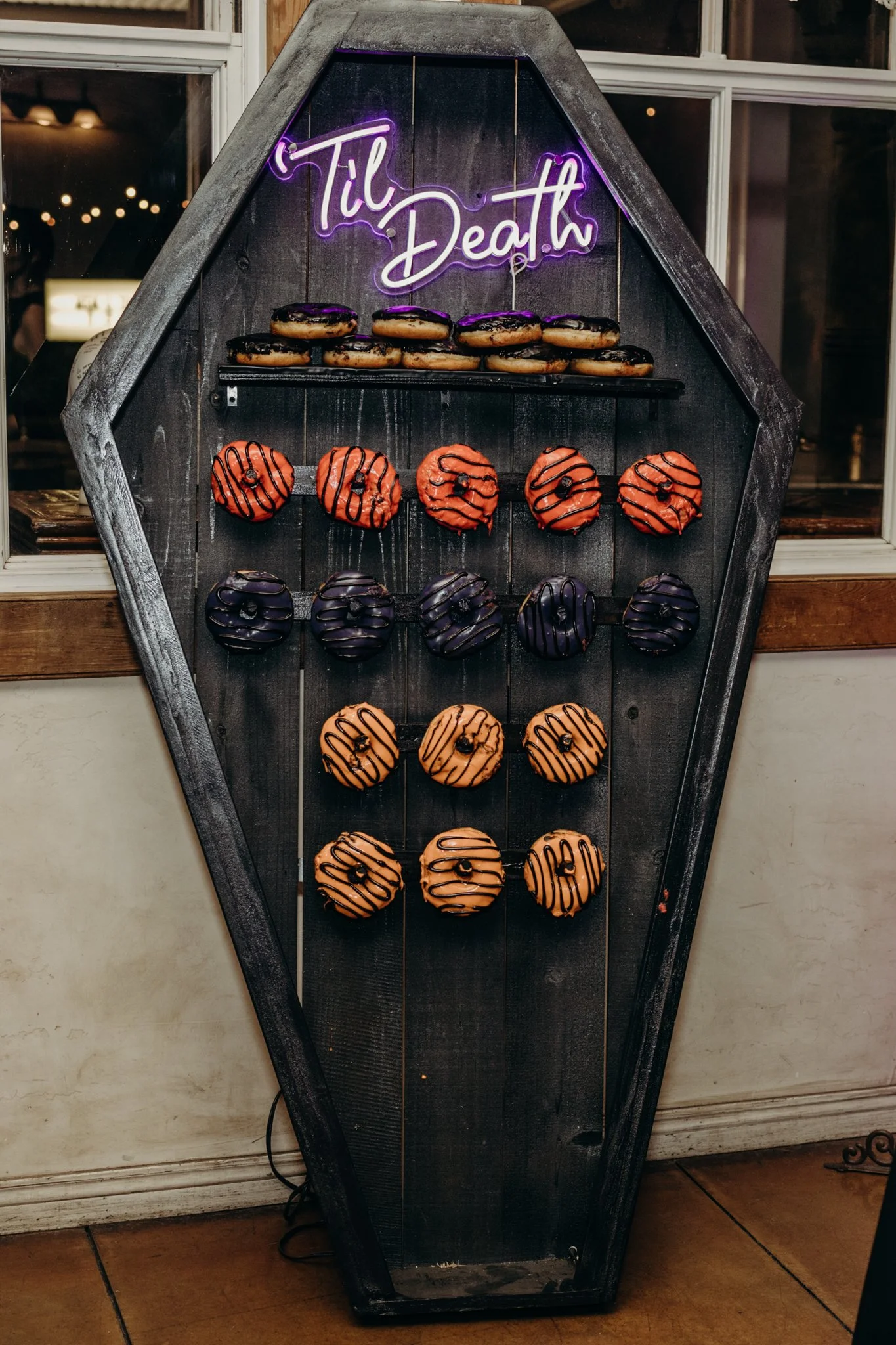 Coffin-shaped donut wall with orange, purple, and black frosted donuts beneath a glowing "'Til Death" neon sign at a Halloween wedding reception.