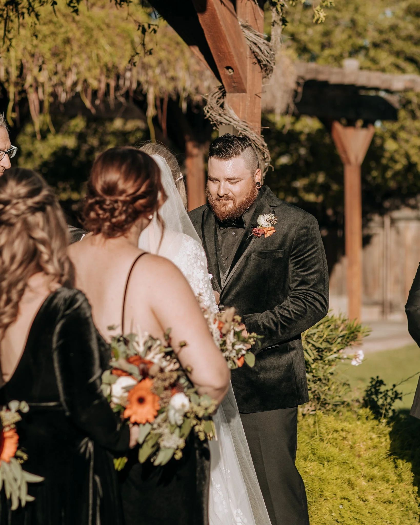 Bride and groom exchanging vows beneath a vine-covered pergola during an outdoor Halloween-themed wedding ceremony with bridesmaids and groomsmen standing beside them.