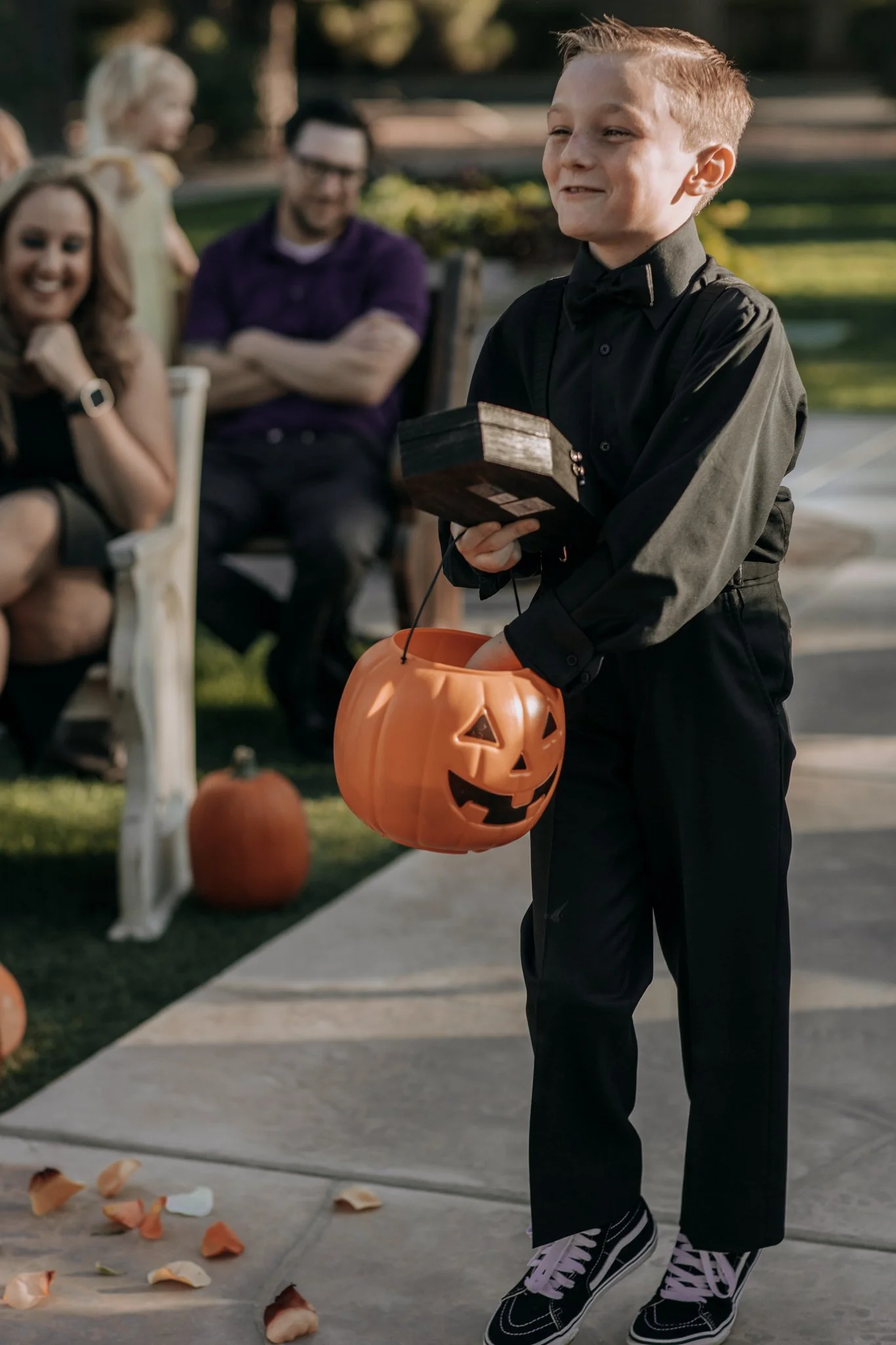 Young ring bearer carrying a ring box and a pumpkin bucket while walking down the aisle during a Halloween-themed outdoor wedding ceremony.