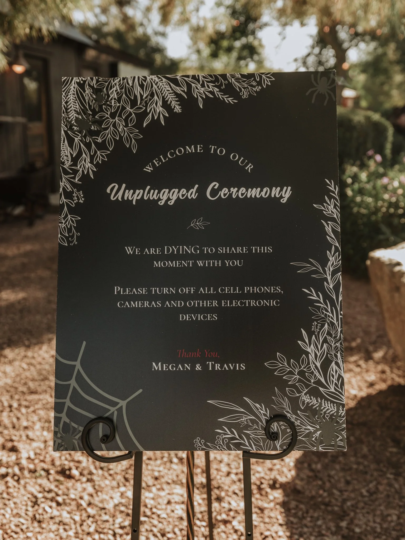 Black unplugged ceremony sign with botanical and spider web illustrations asking guests to put away phones and cameras during the wedding ceremony.