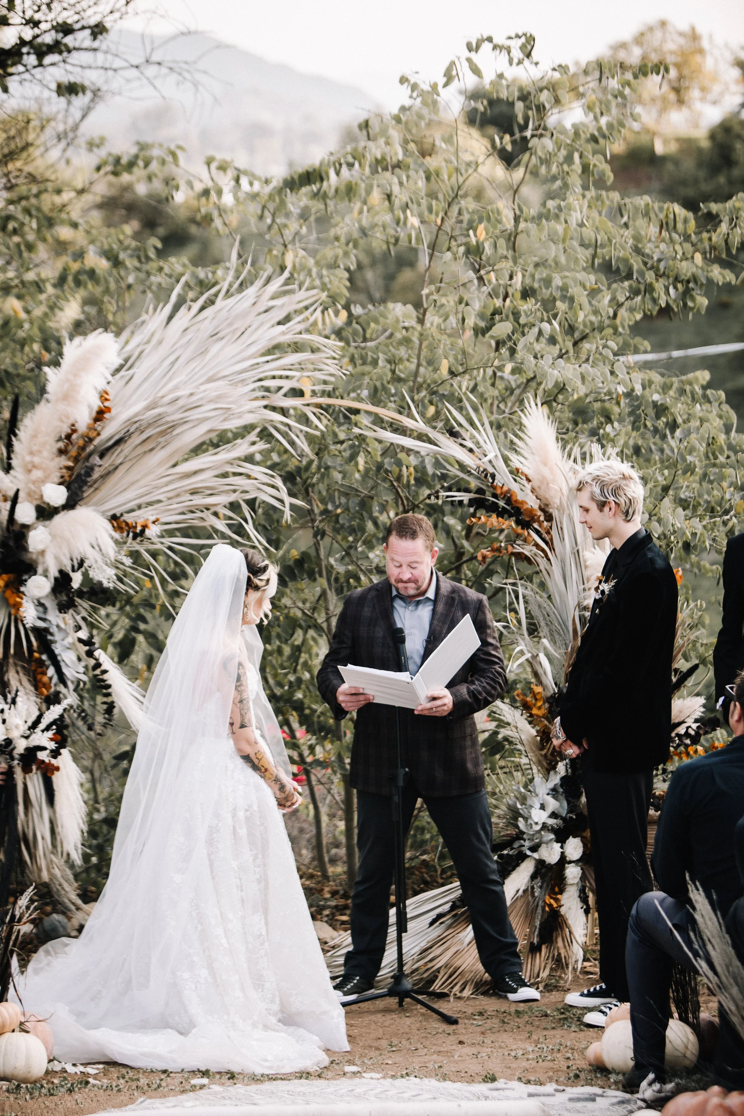 Bride and groom stand beneath a dramatic dried floral ceremony arch with pampas grass, white florals, and black accents during their outdoor Halloween wedding.