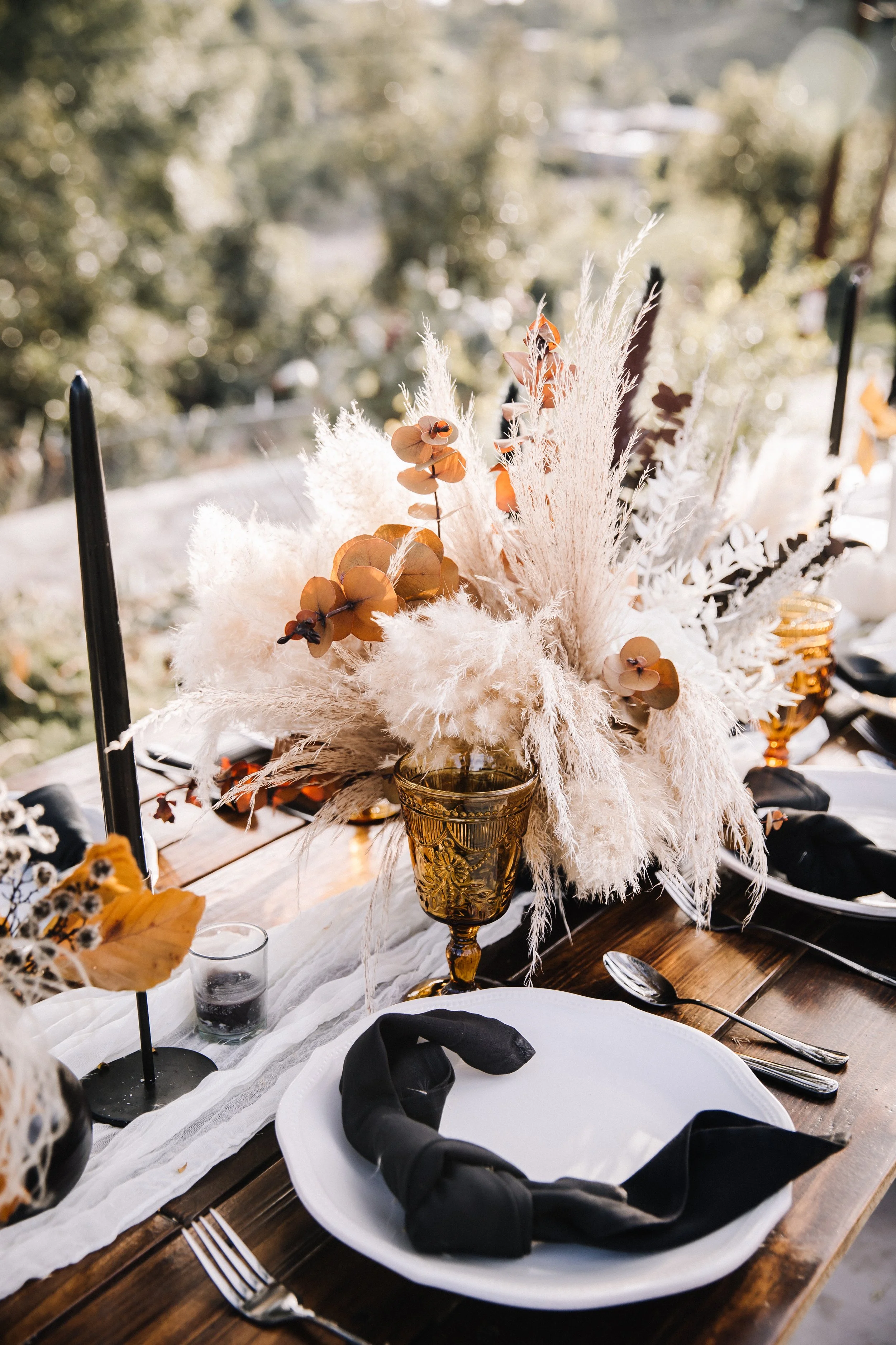 Dark romantic wedding theme centerpiece with pampas grass, orchids, dried florals, black foliage, and warm autumn tones.