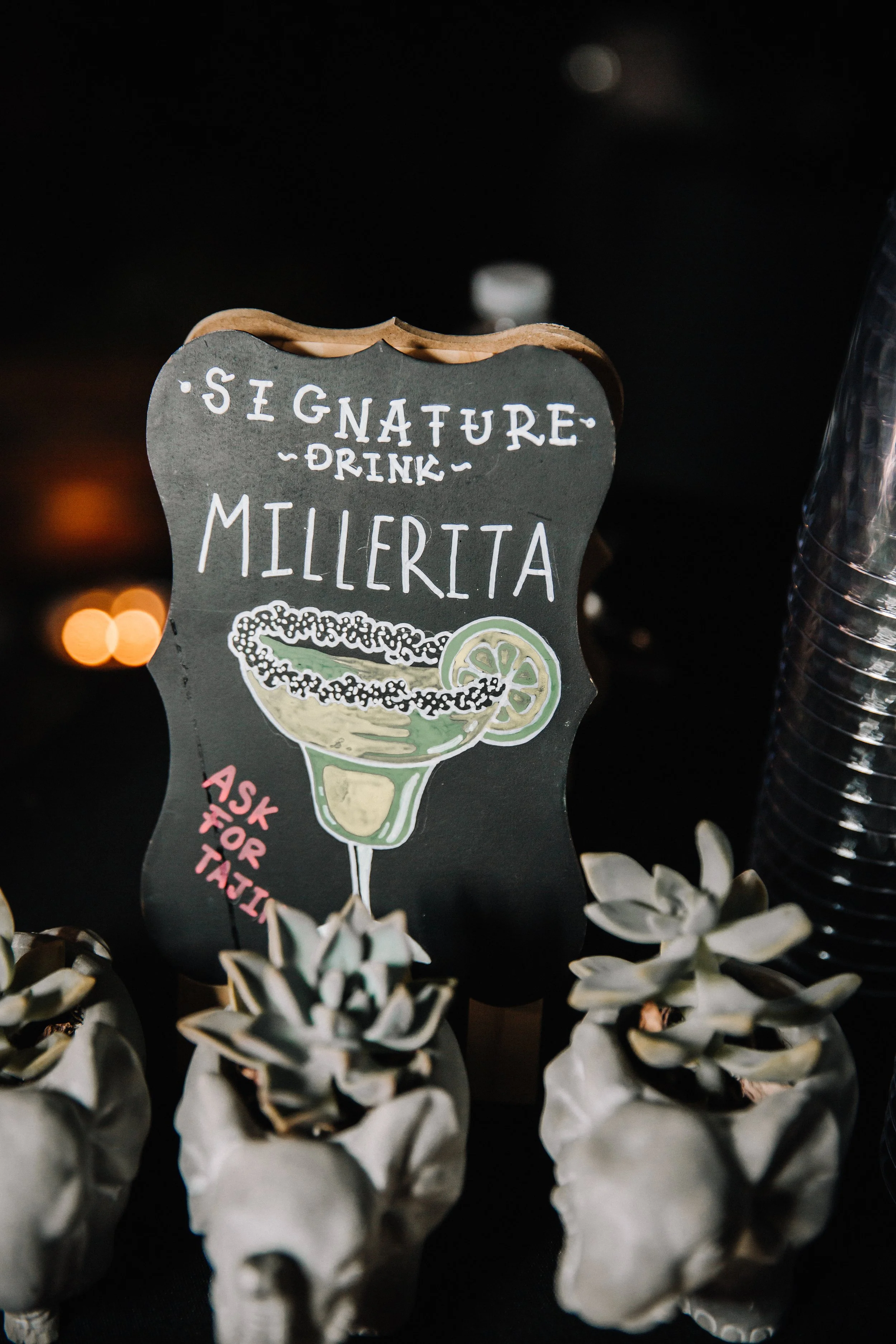 Hand-painted signature cocktail sign with skull planters and playful Halloween wedding details at the reception bar.