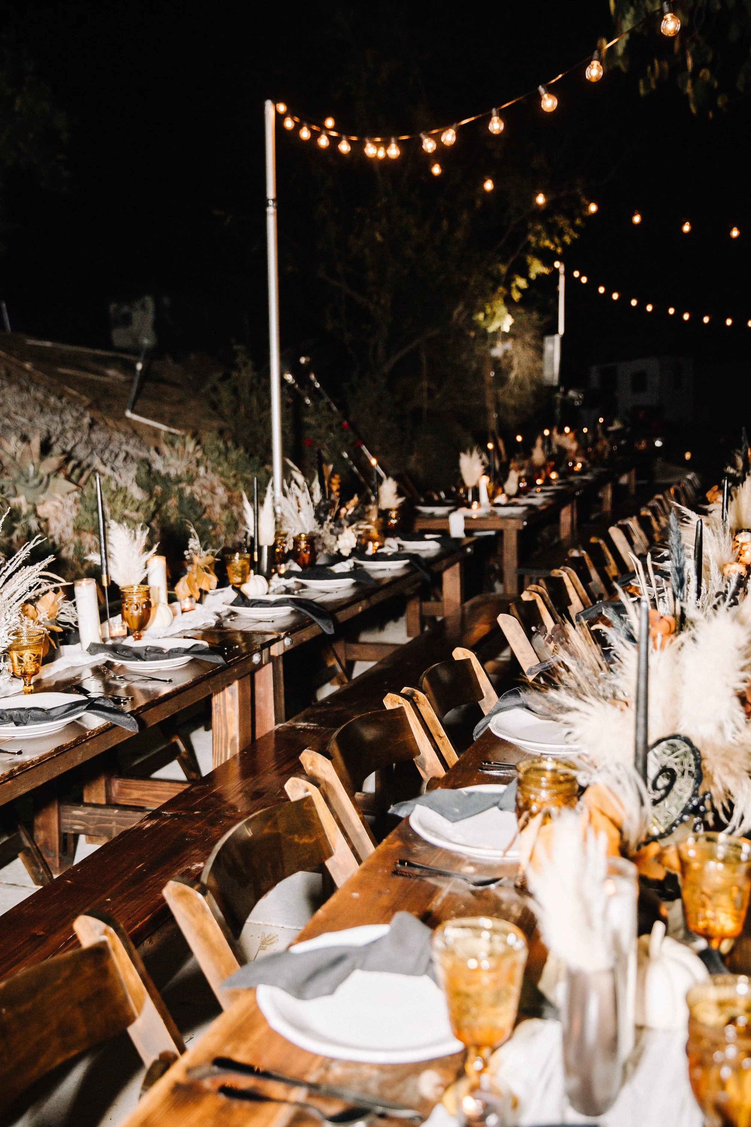 Elegant Halloween wedding reception table with white dinnerware, black napkins, amber goblets, candles, pampas grass, and white pumpkins on a rustic wood table.