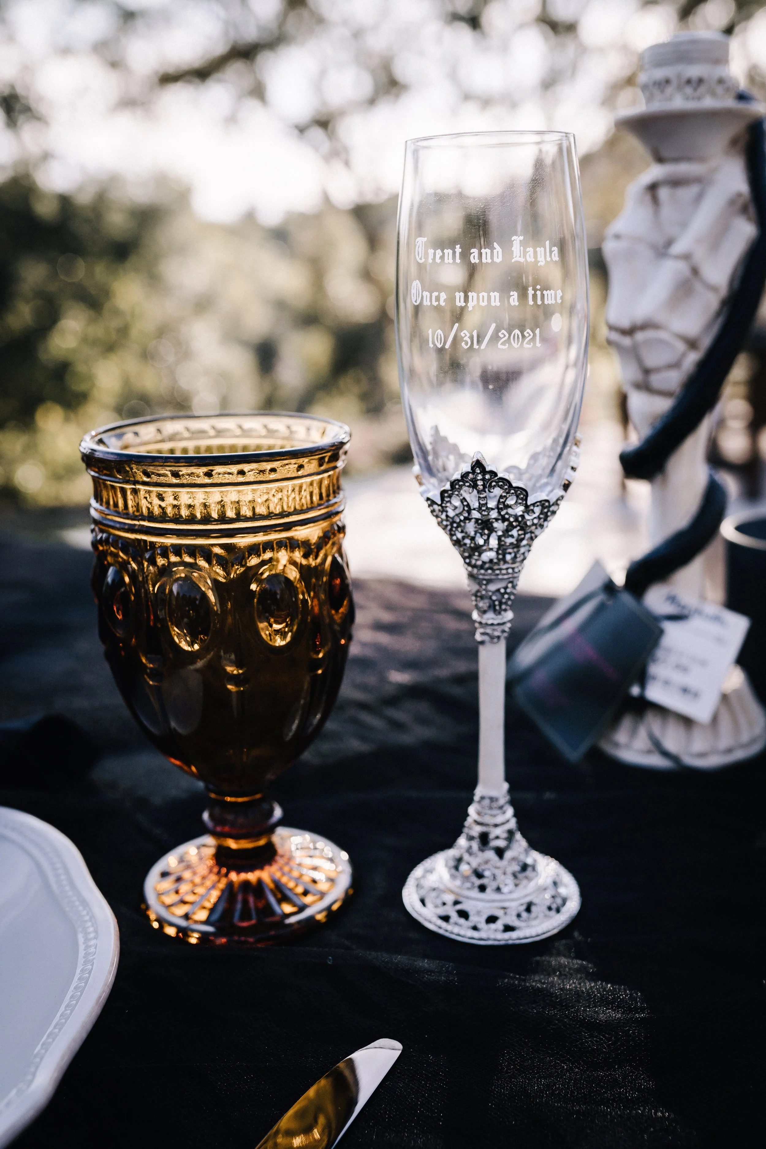 Personalized champagne flute engraved with the couple's names and wedding date beside an amber goblet on a dark romantic Halloween wedding tablescape.
