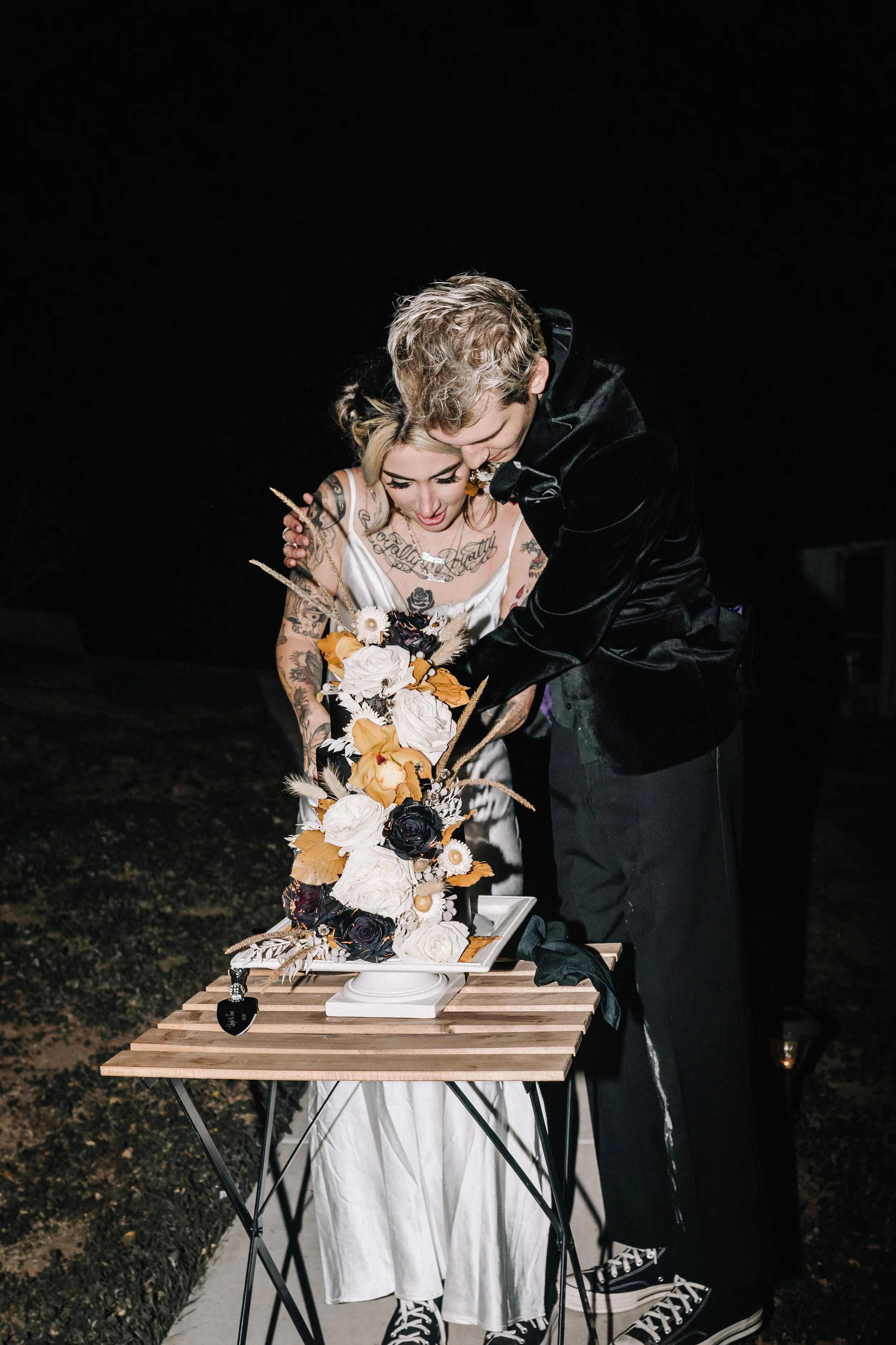 Bride and groom cut a dramatic floral-covered wedding cake during their outdoor Halloween wedding reception under the night sky.