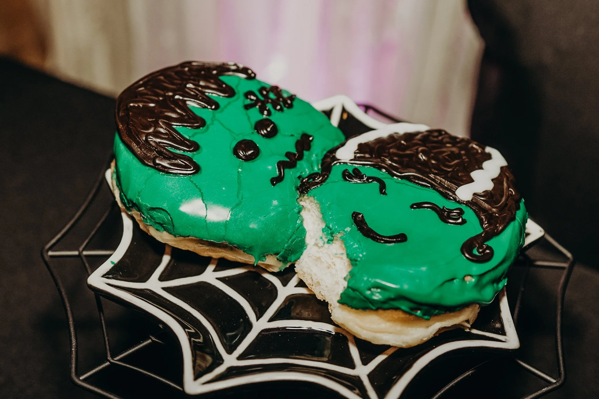 Frankenstein-inspired green frosted donut with a bite taken out, displayed on a black spiderweb plate as a fun Halloween wedding dessert.
