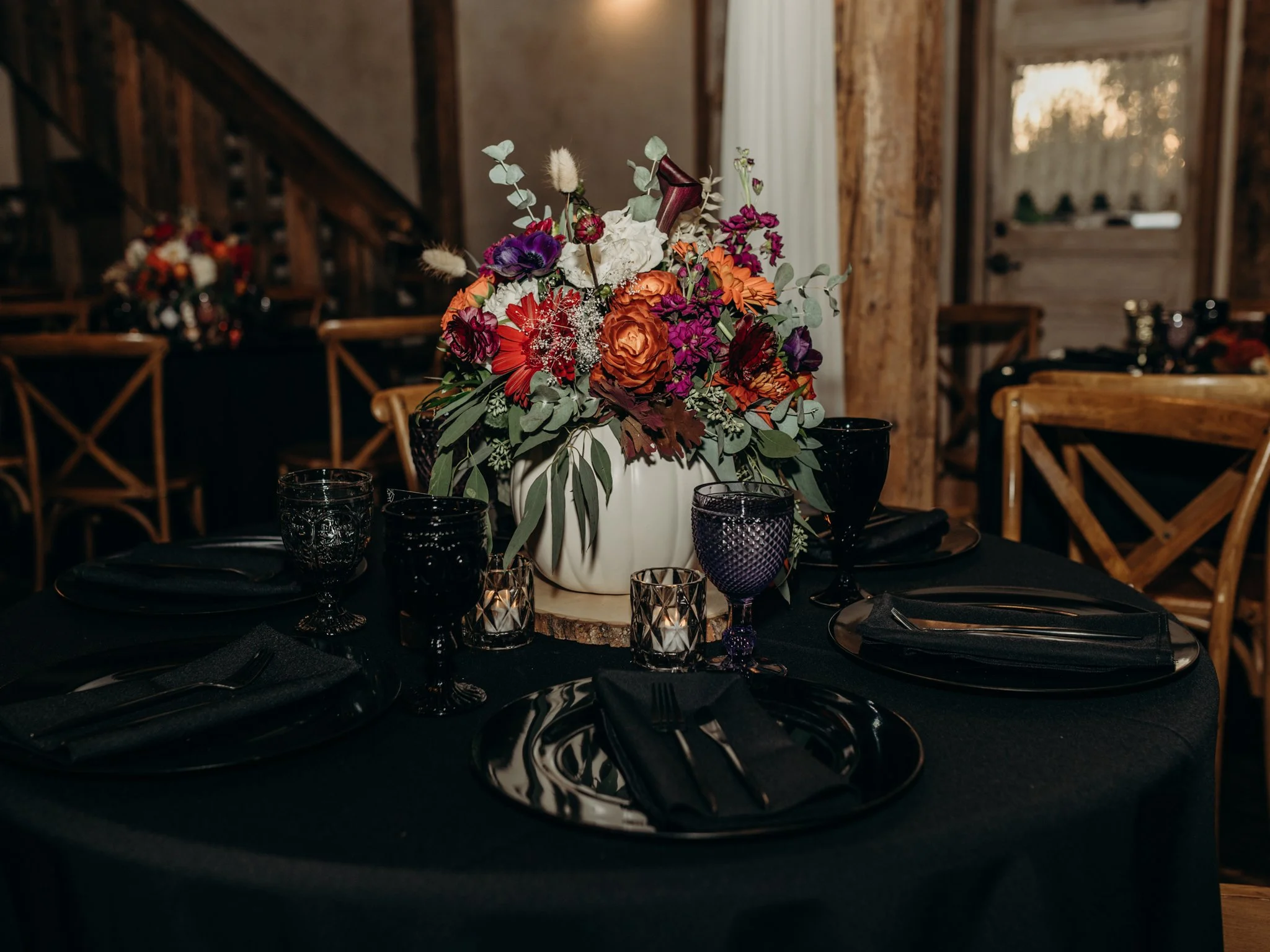 Moody Halloween wedding reception table with black linens, black dinnerware, dark glassware, and a vibrant autumn floral centerpiece featuring orange, burgundy, purple, and white blooms.
