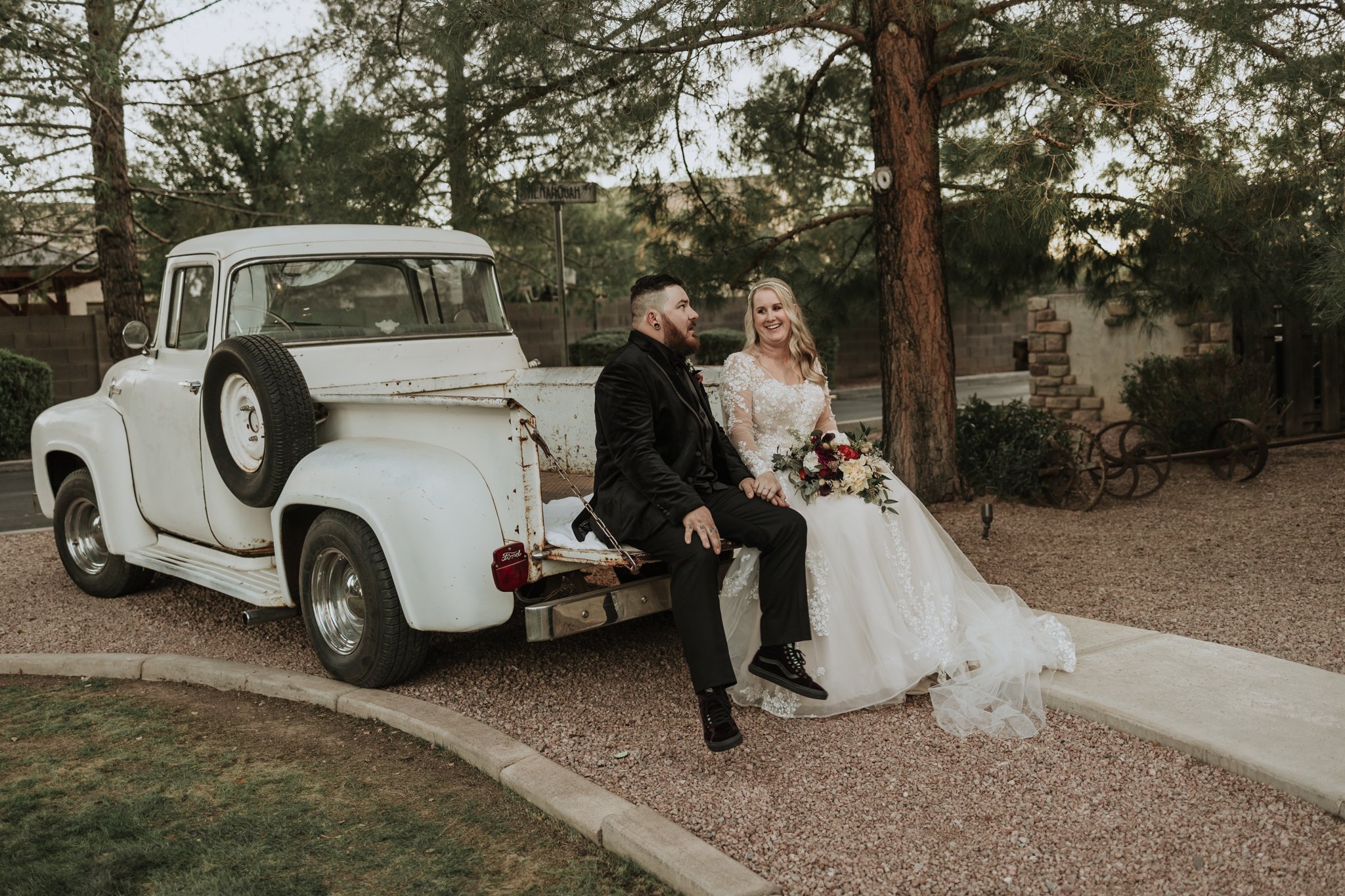 Bride and groom sitting together on the tailgate of a vintage white pickup truck during relaxed outdoor wedding portraits.