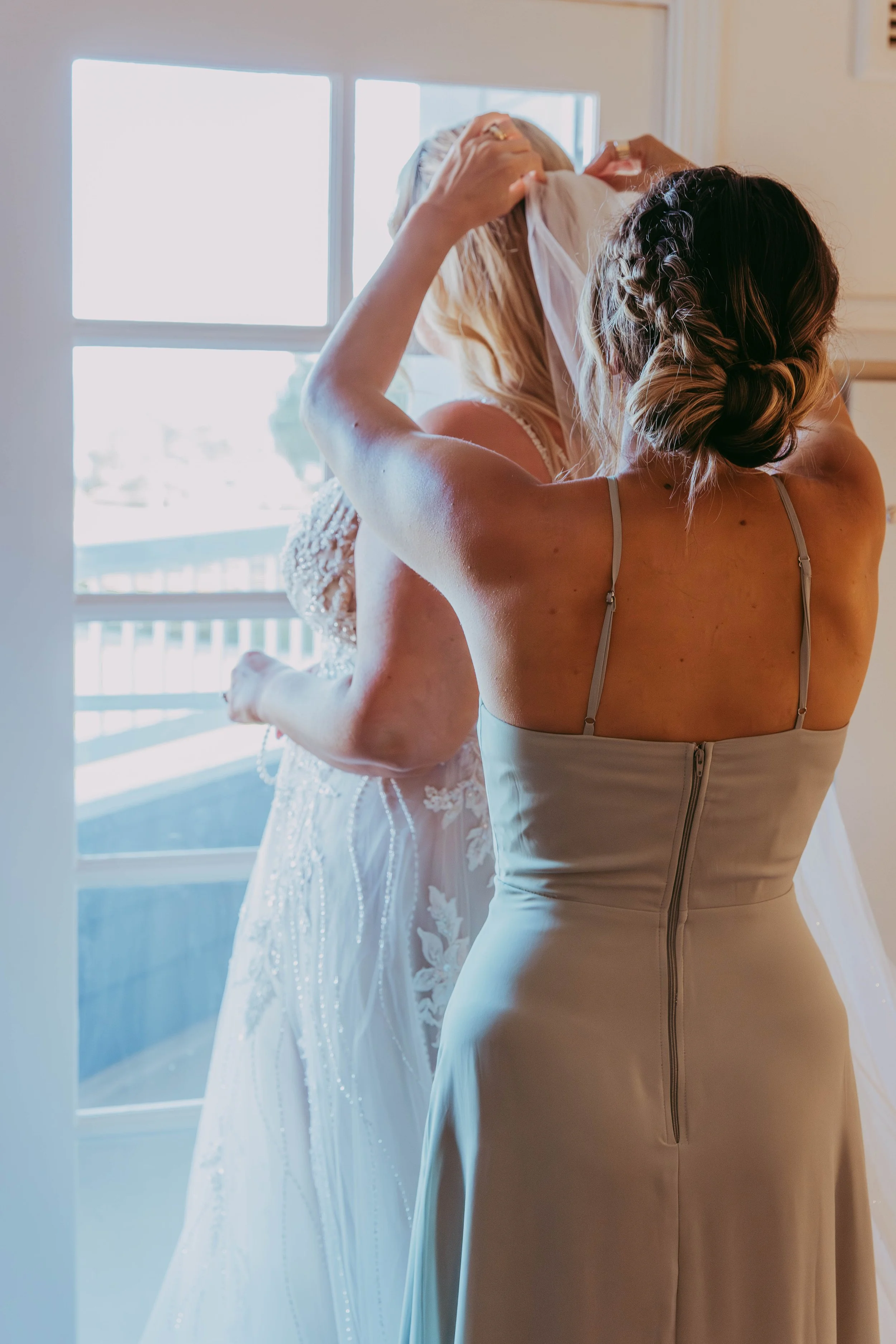 Bridesmaid helps the bride with her veil by a harbor-view window before a Dana Point wedding