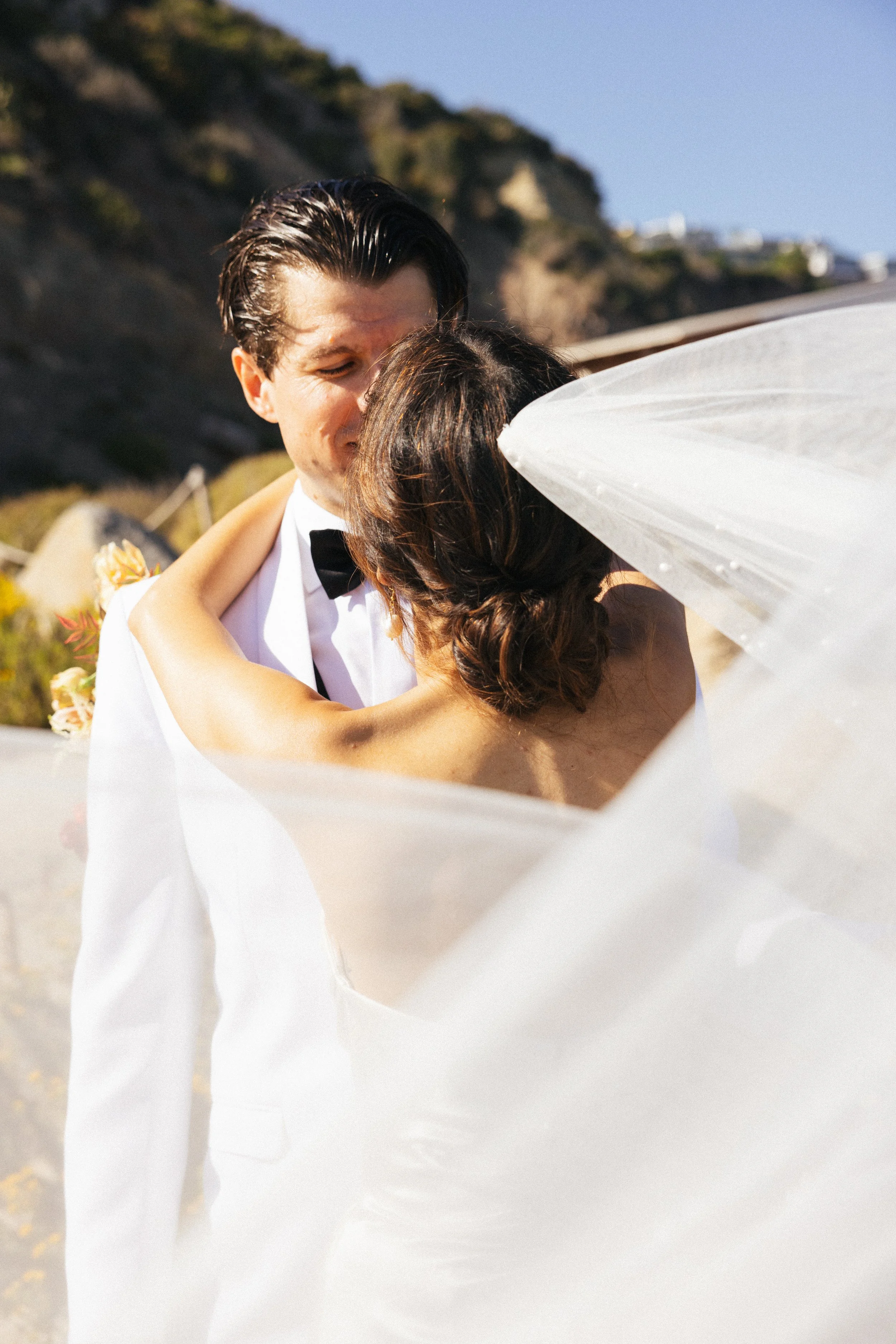Bride and groom embrace as her veil lifts in the wind on the Dana Point coast