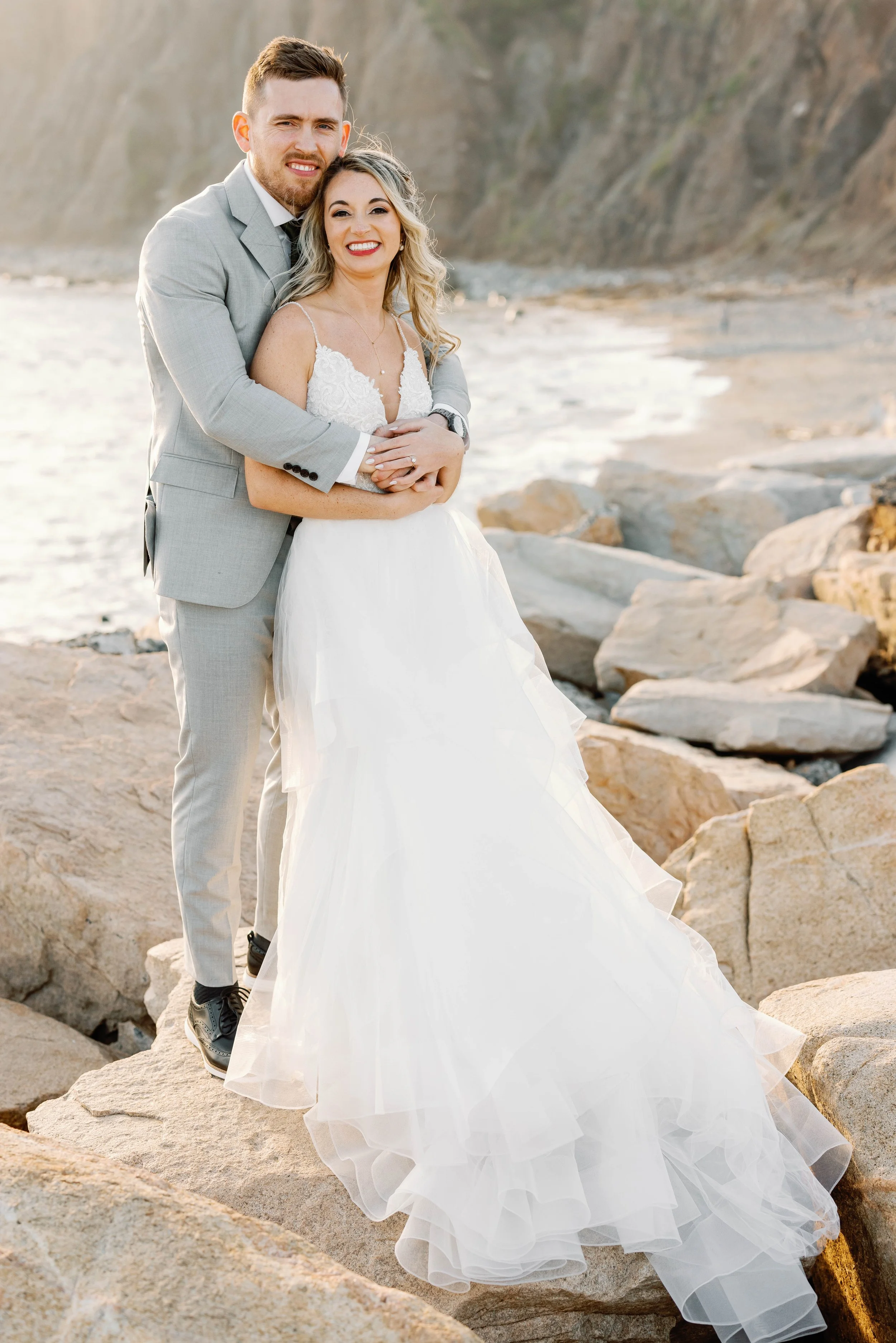 Bride and groom embrace on the rocks along the Dana Point coast at golden hour