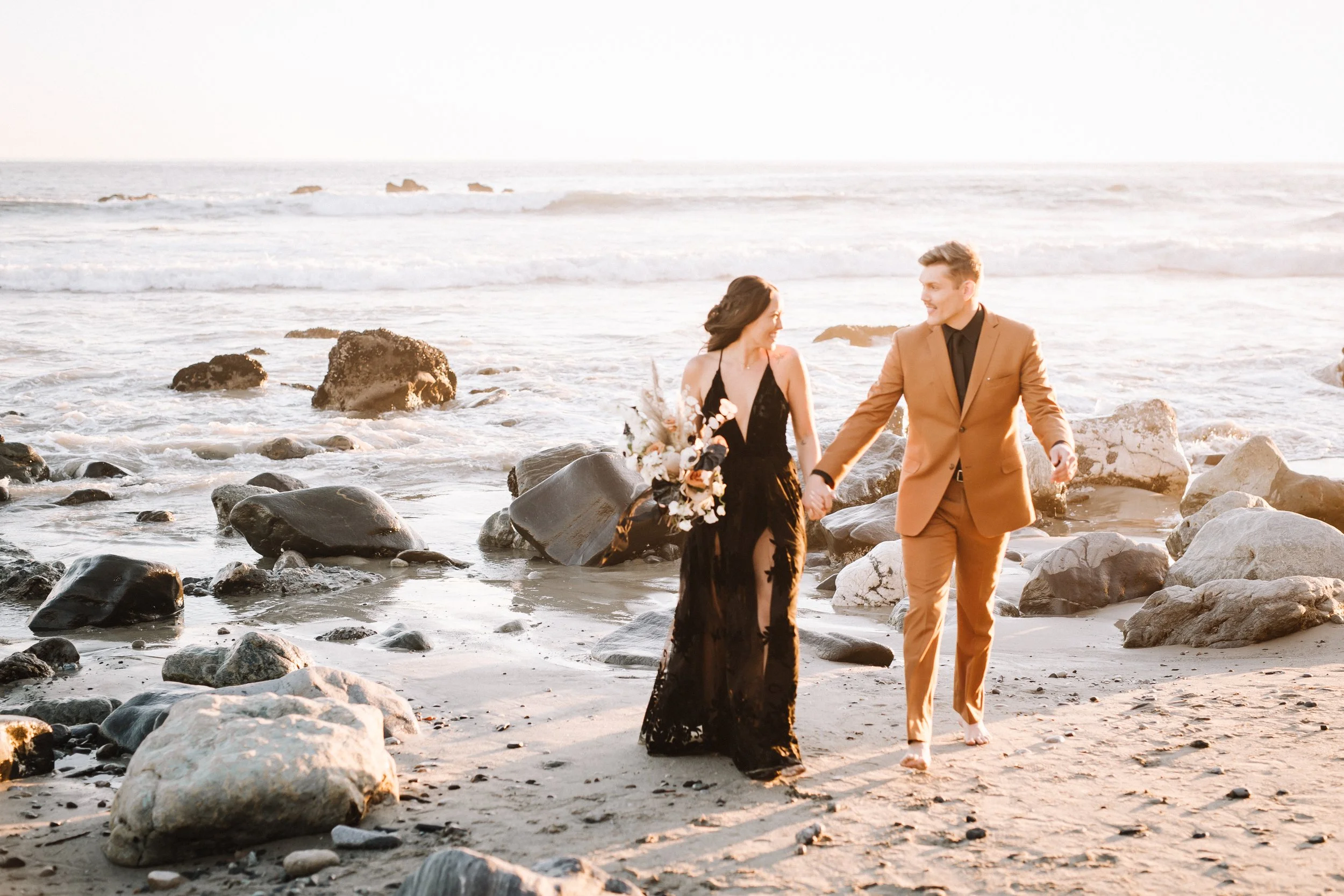 Couple walk the rocky shoreline at a Dana Point beach wedding, bride in a black gown