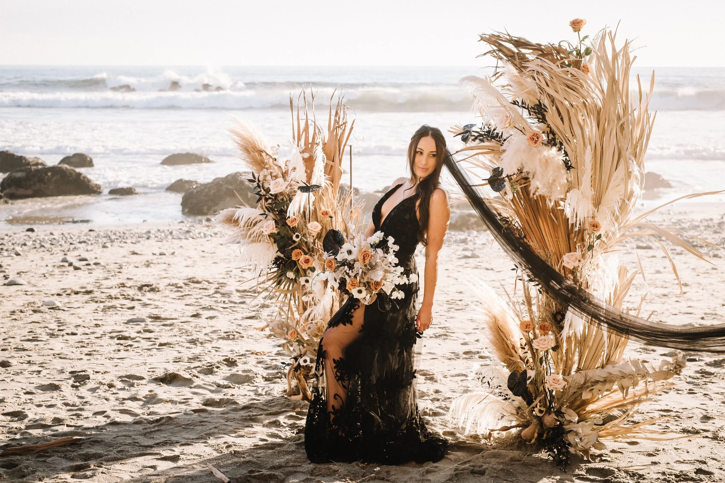 Bride in a black lace gown between two dried palm installations on the sand at a Dana Point beach wedding