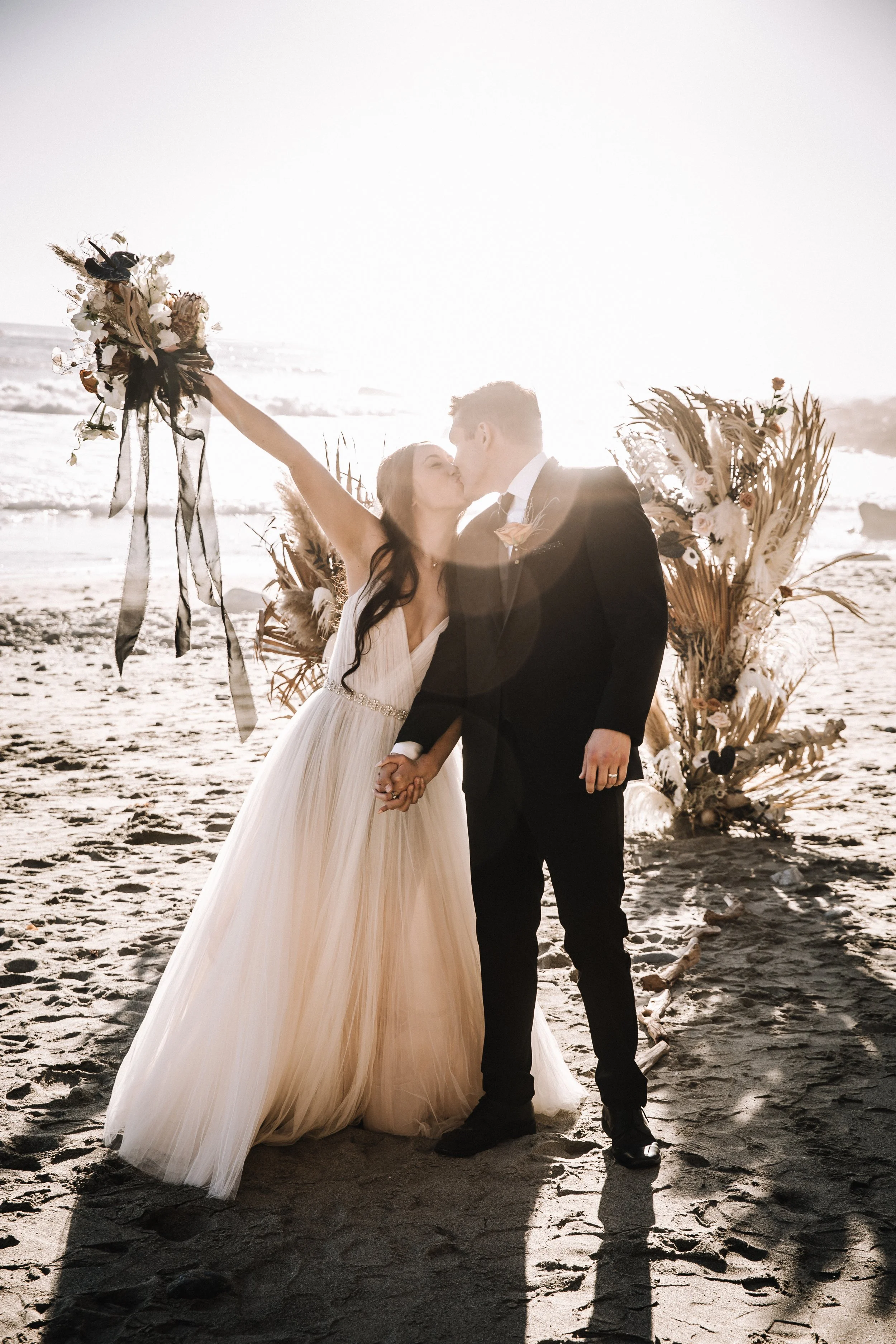 Couple kiss as the bride raises a bouquet at a Dana Point beach wedding ceremony