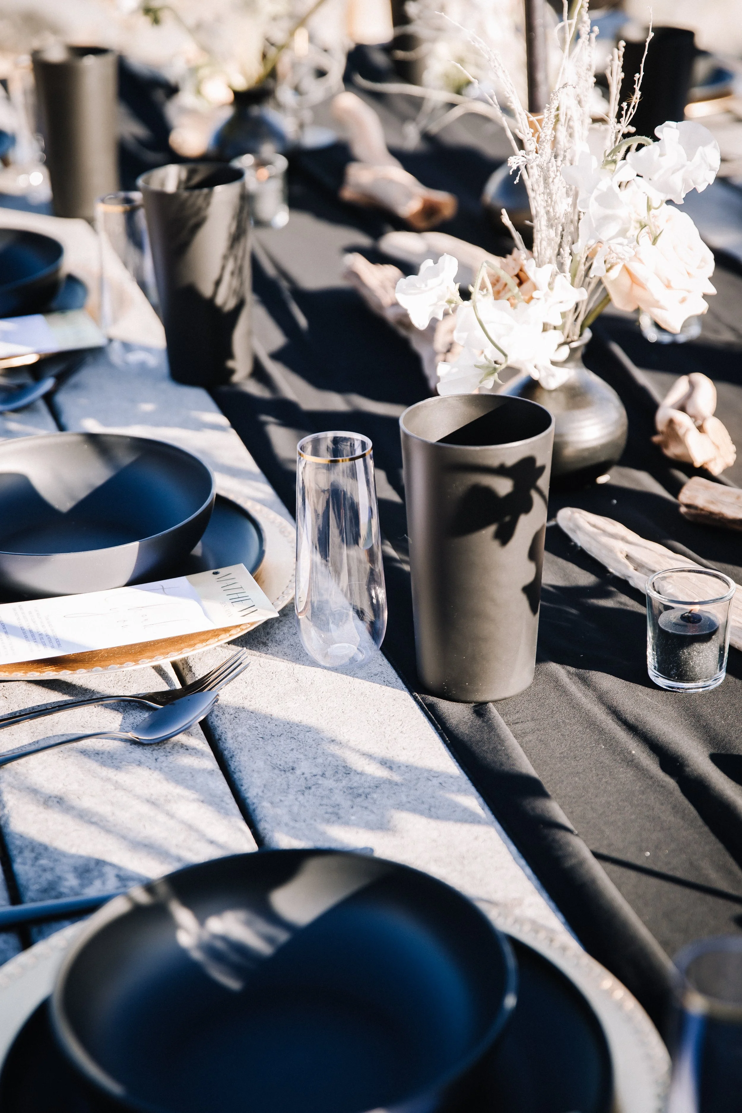 Black place settings, gold-rim glassware and driftwood on a black table at a beach wedding