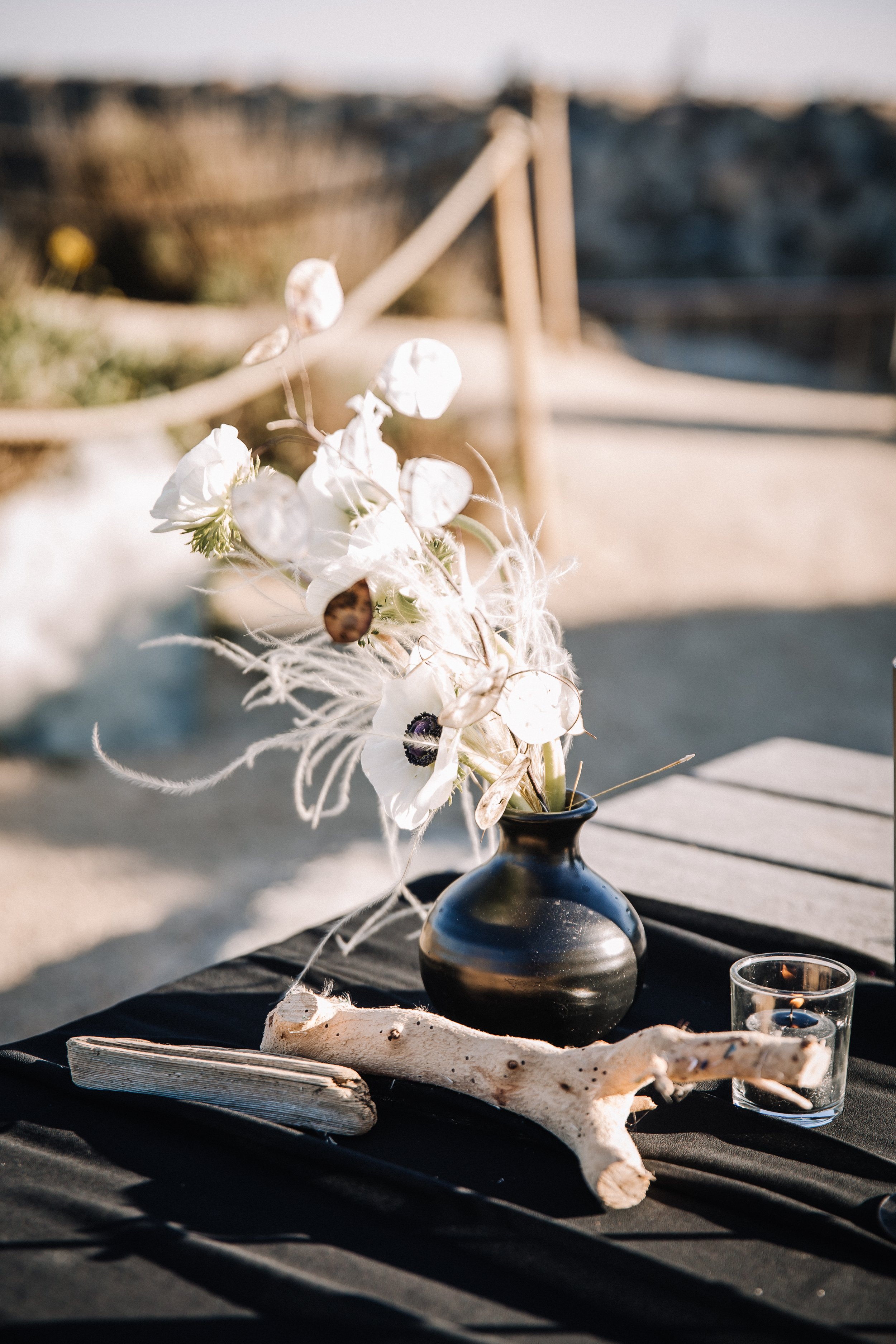 White anemones and lunaria in a black bud vase on driftwood at a beach wedding