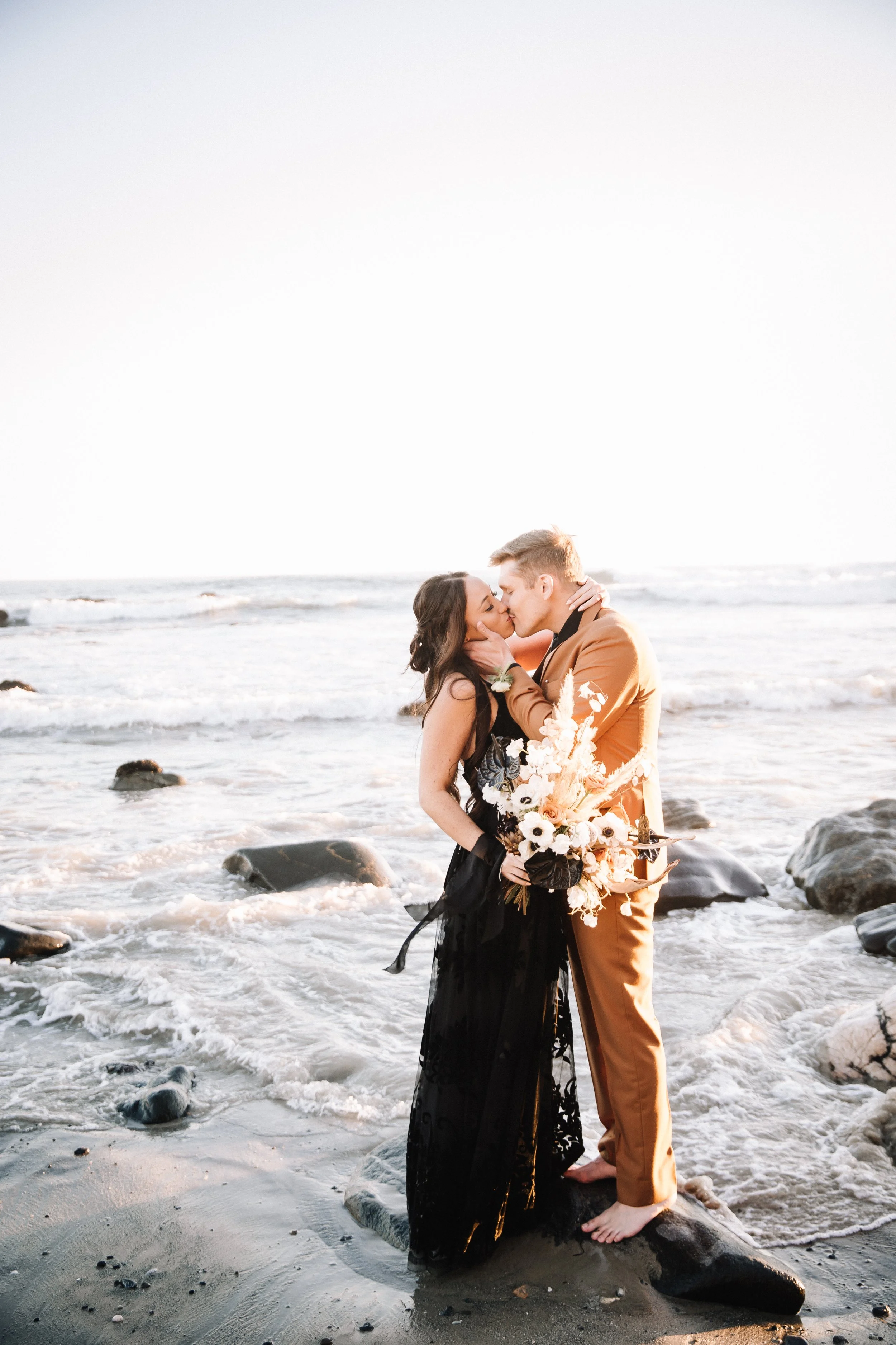 Couple on a rock in the surf at an alternative Dana Point beach wedding, groom in a caramel suit