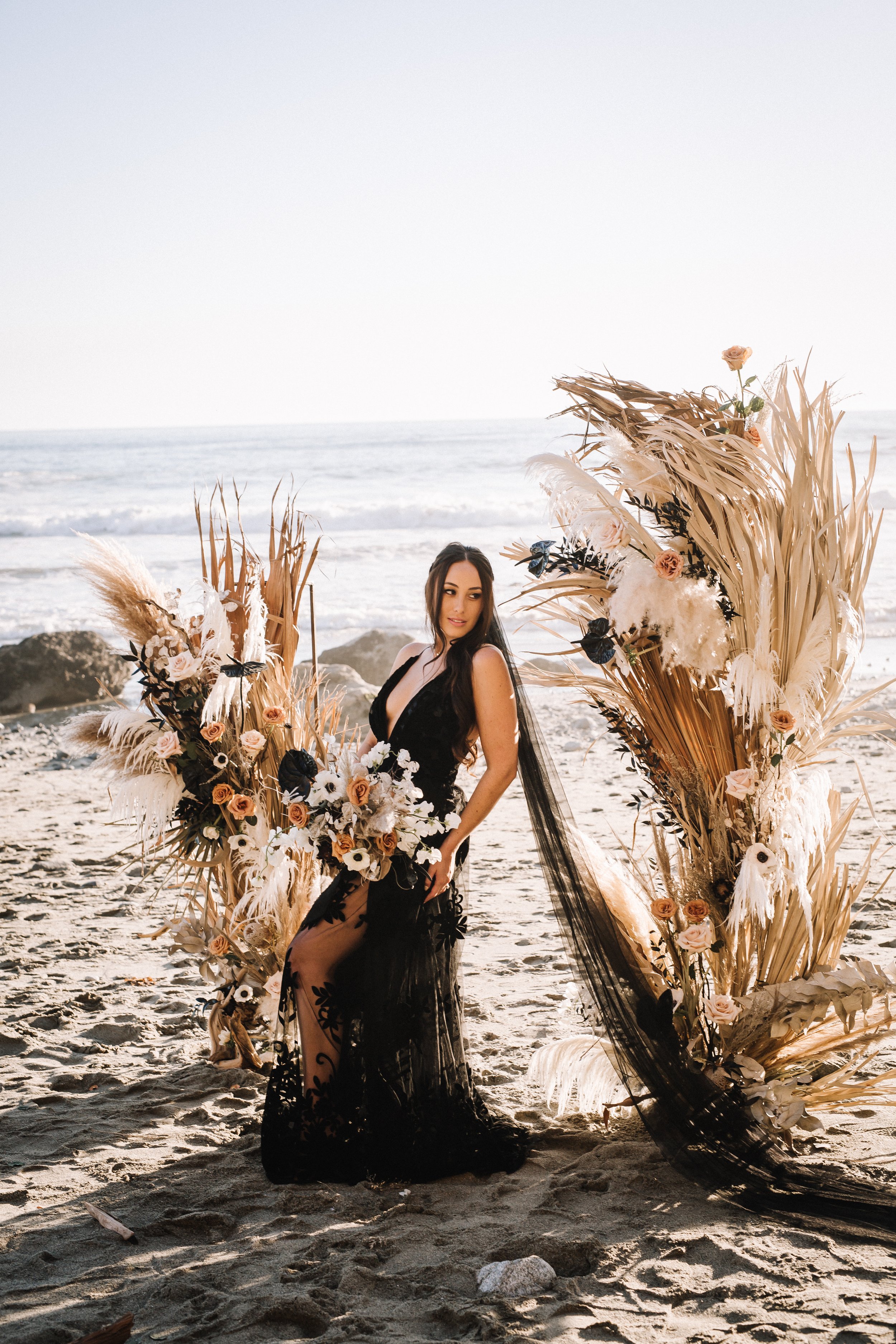 Bride in a black lace gown between dried palm and pampas installations at a Dana Point beach wedding