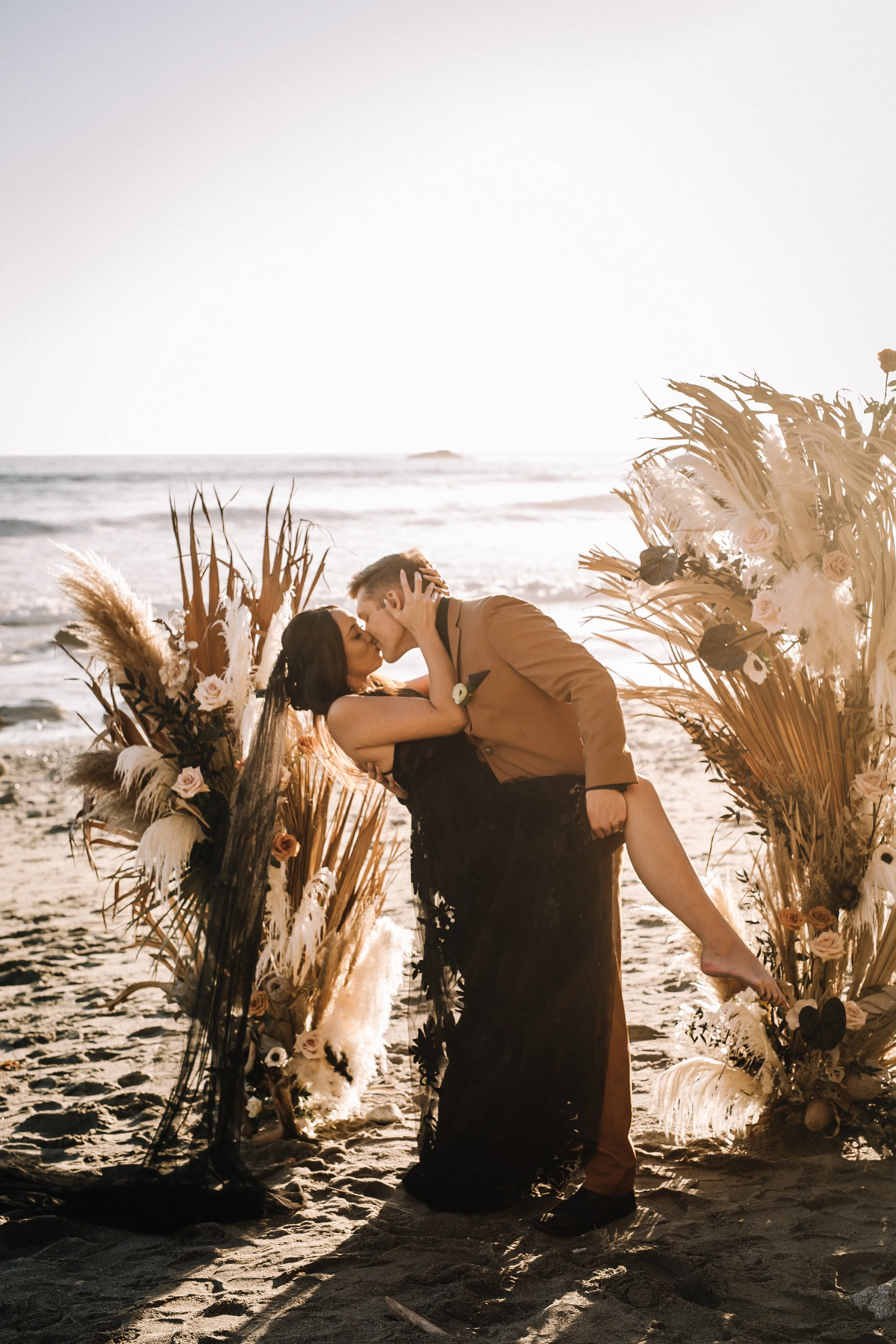 Couple kissing on a rock in the surf, groom in a caramel suit, at a Dana Point beach wedding