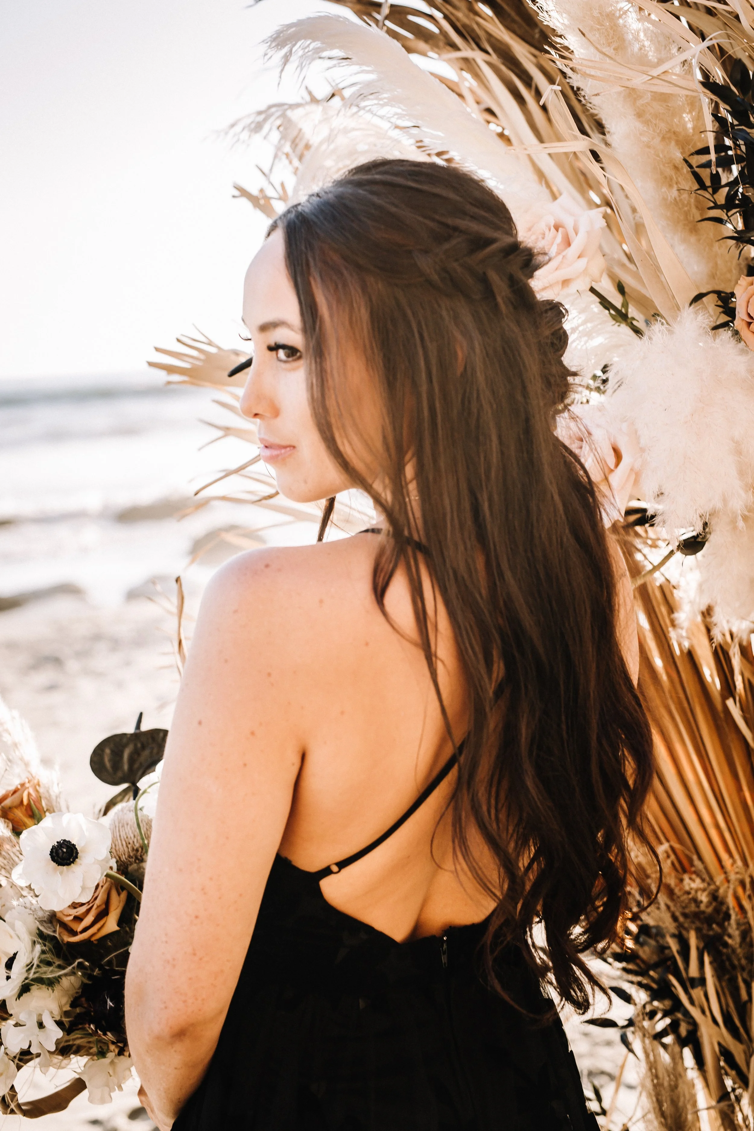 Bride in a black lace gown looks over her shoulder beside a dried palm installation at a Dana Point beach wedding
