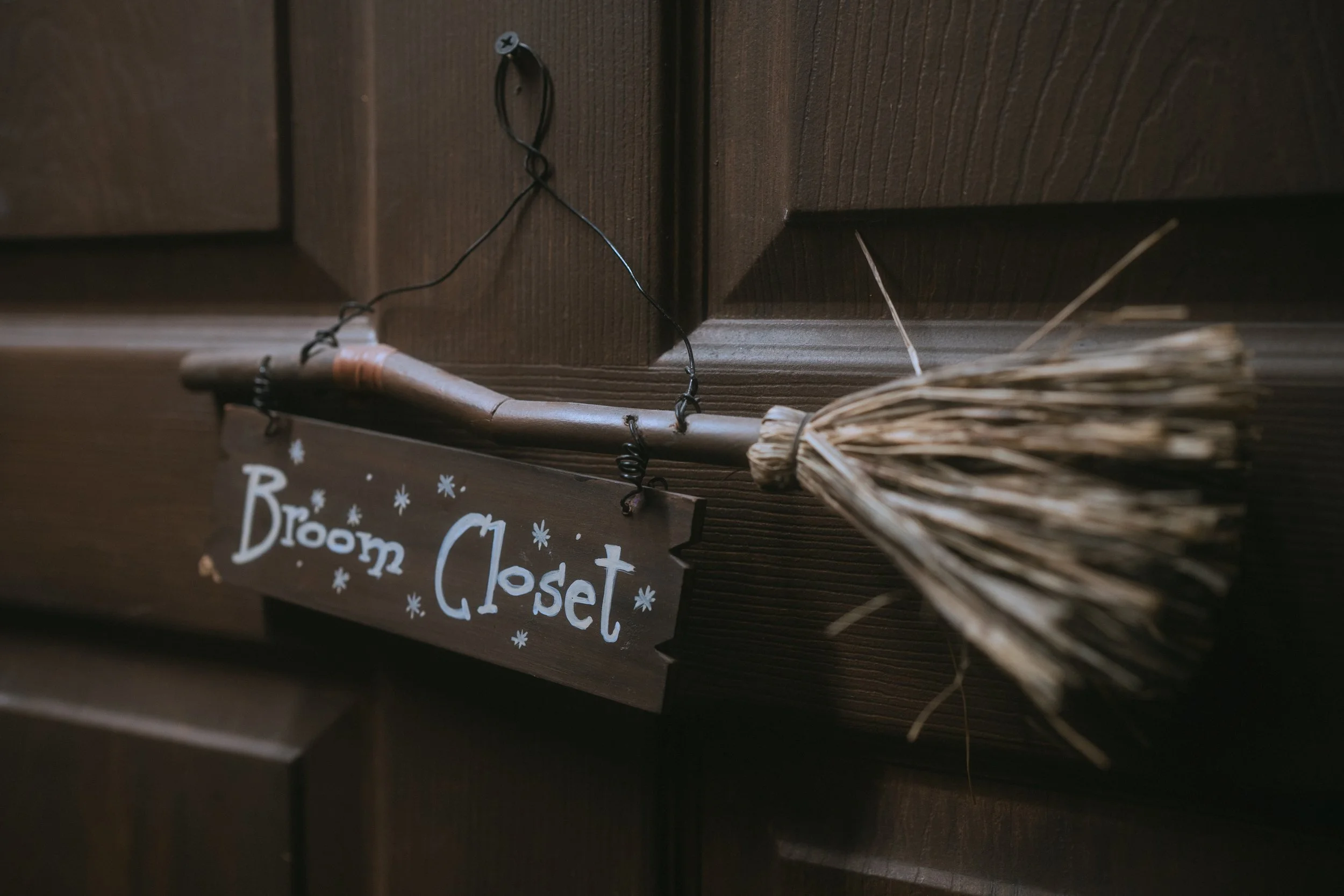 Gothic “Broom Closet” sign hanging from a wooden broom on a dark wood wall as part of Halloween-themed wedding decor