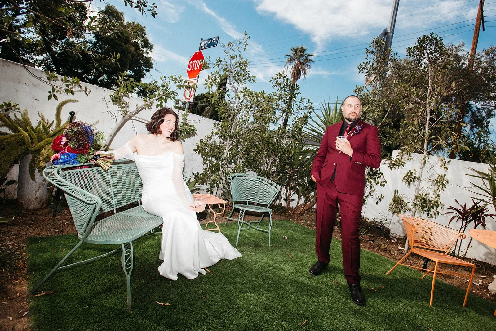 Bride seated on a mint green vintage bench and groom standing nearby with a cocktail in an editorial-style wedding portrait set in a private courtyard.