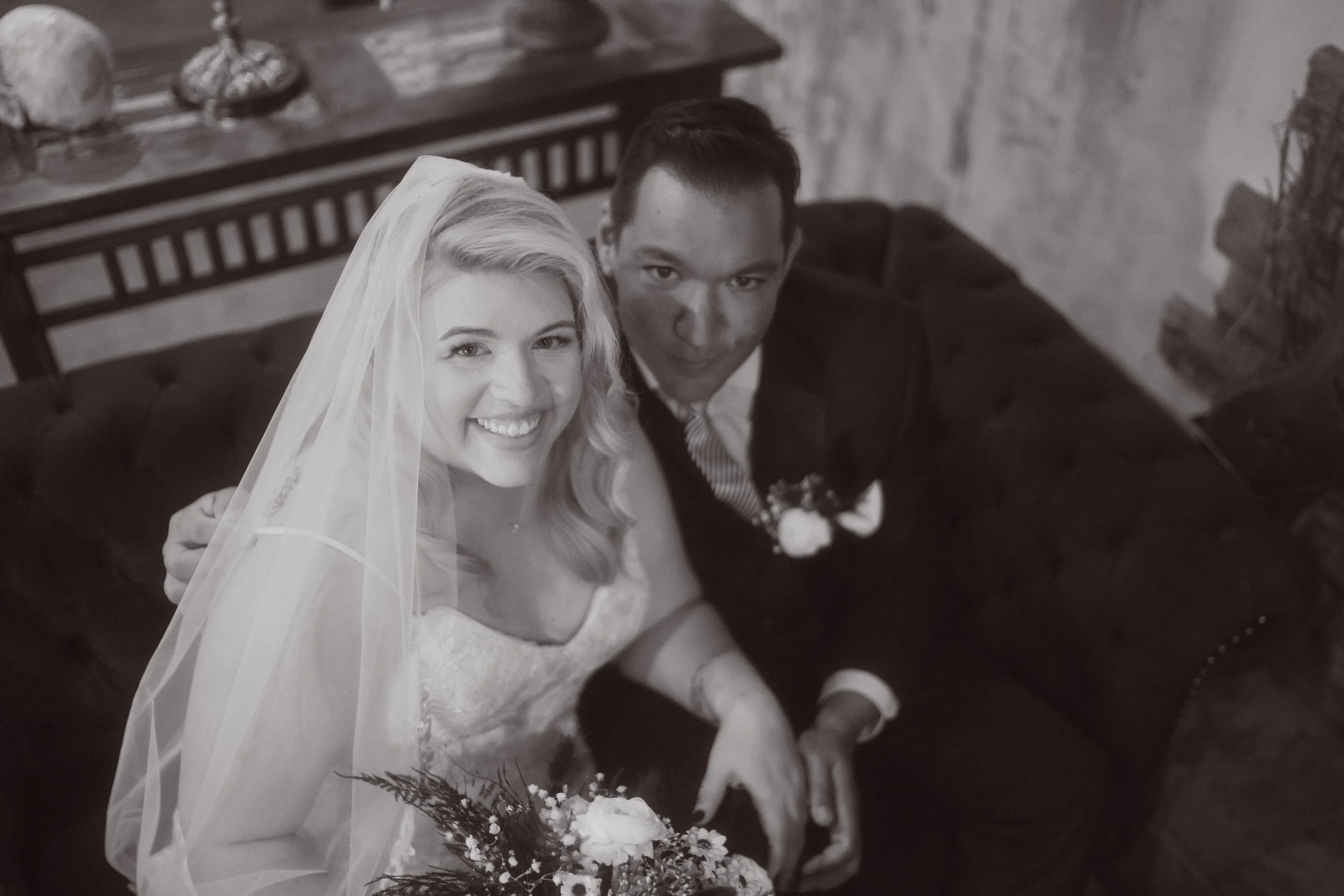 Overhead portrait of bride and groom seated together smiling at the camera, with bride holding bouquet and groom in suit