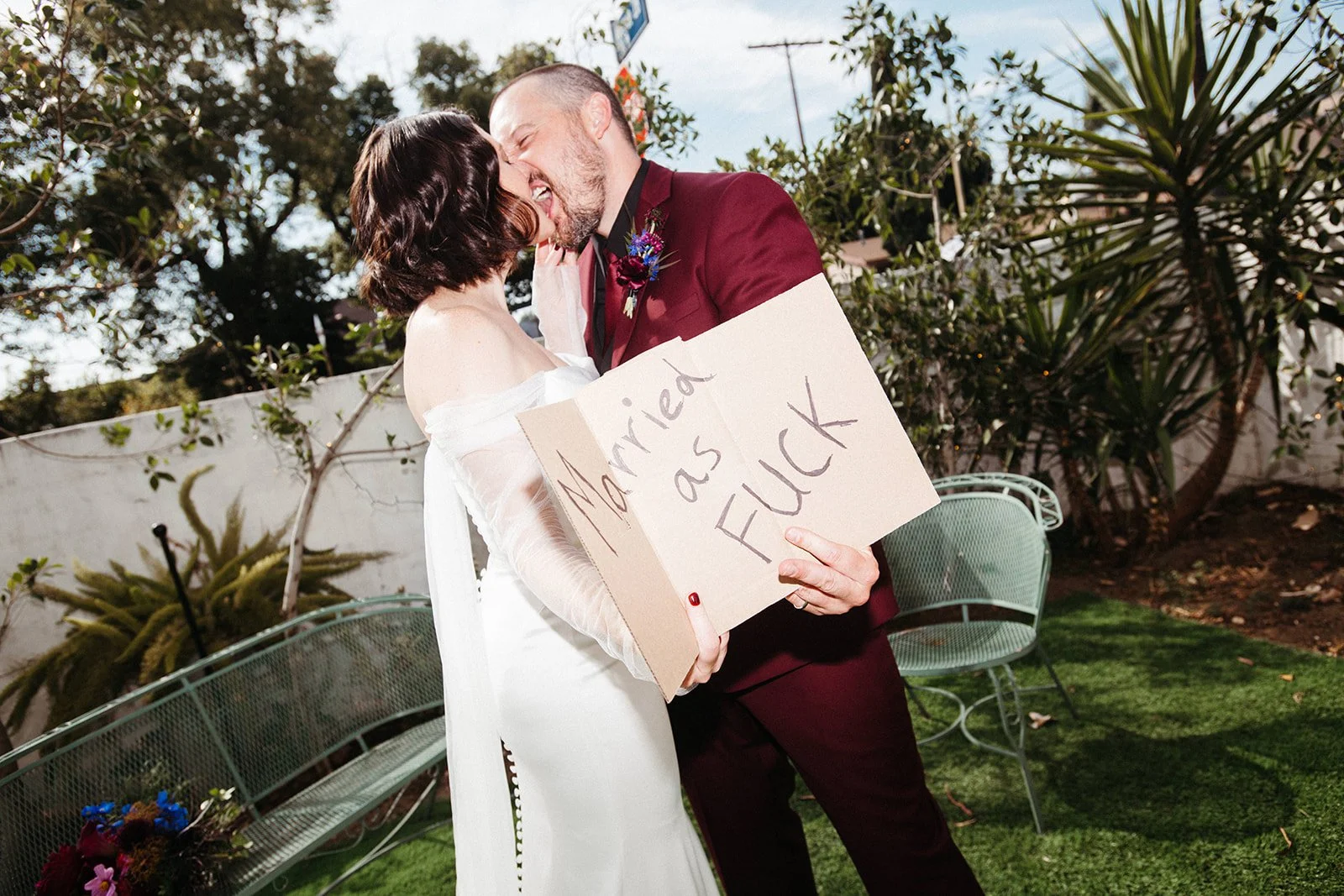 Newlywed couple kisses while holding a playful "Married as Fuck" sign in a garden courtyard during their wedding portrait session.