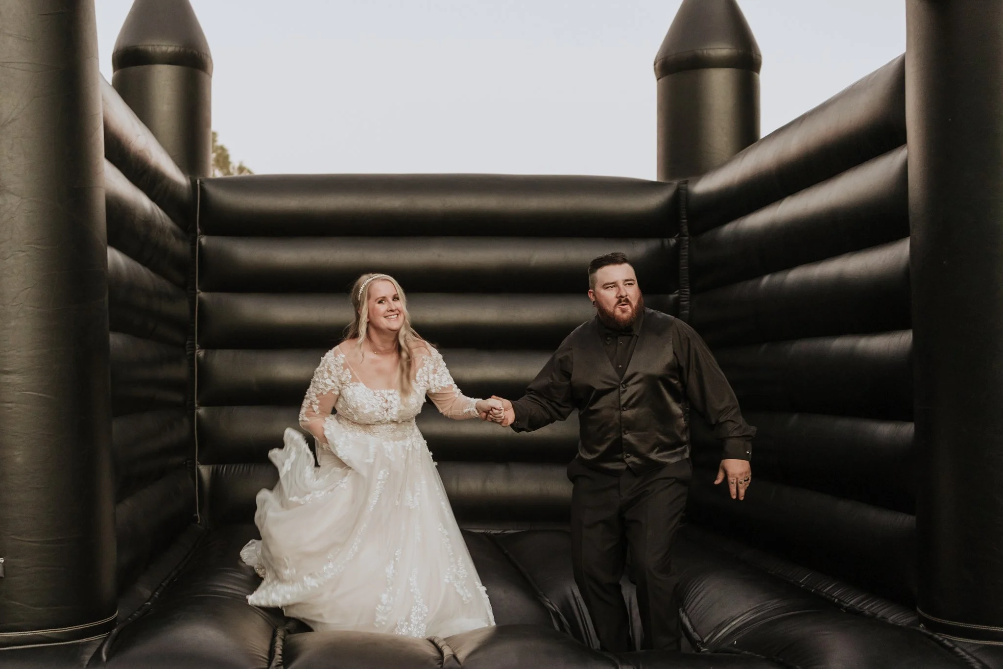 Couple laughing and jumping together inside a black bounce house during the outdoor wedding reception entertainment.