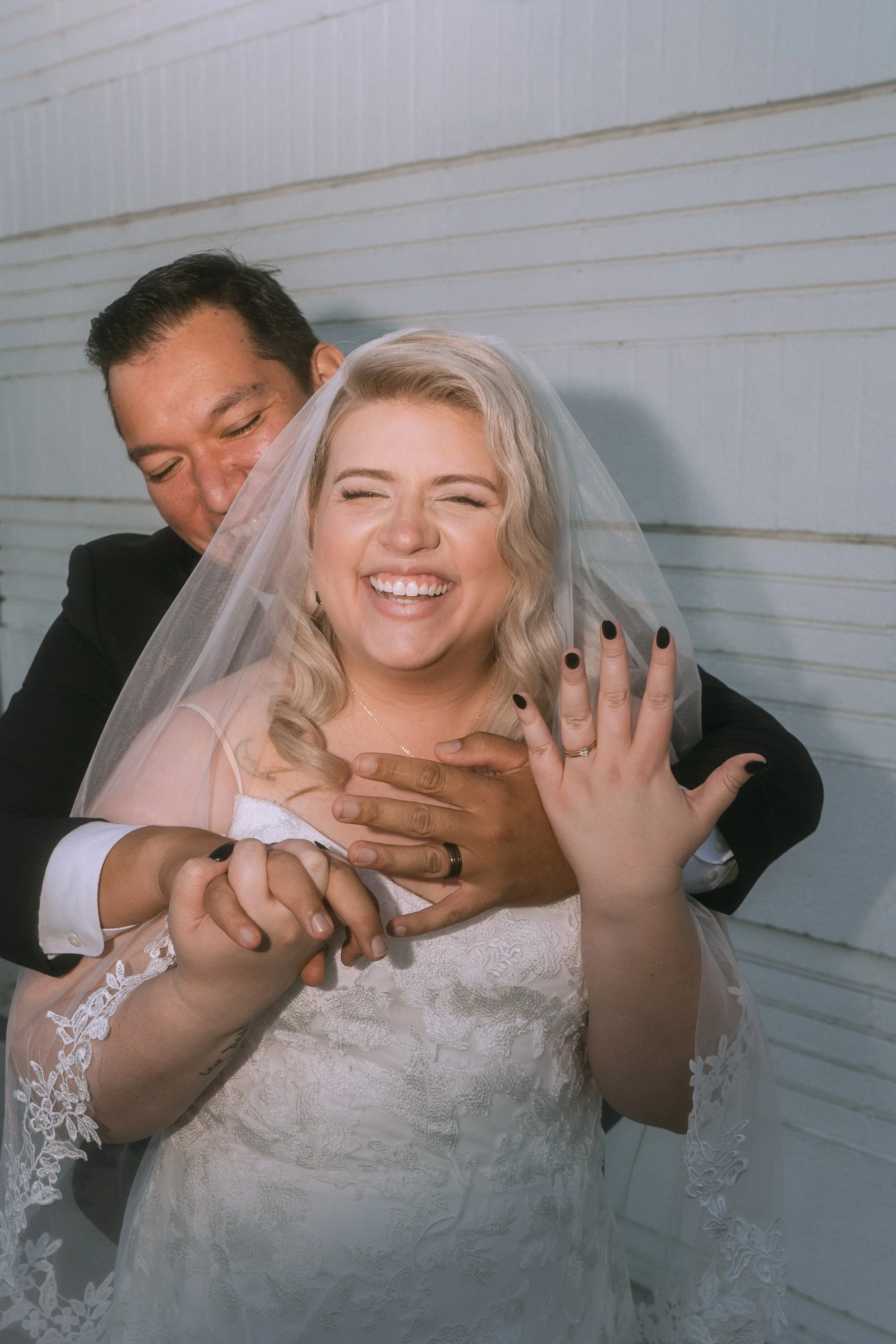 Bride smiling and showing wedding ring while groom embraces her from behind during joyful wedding moment